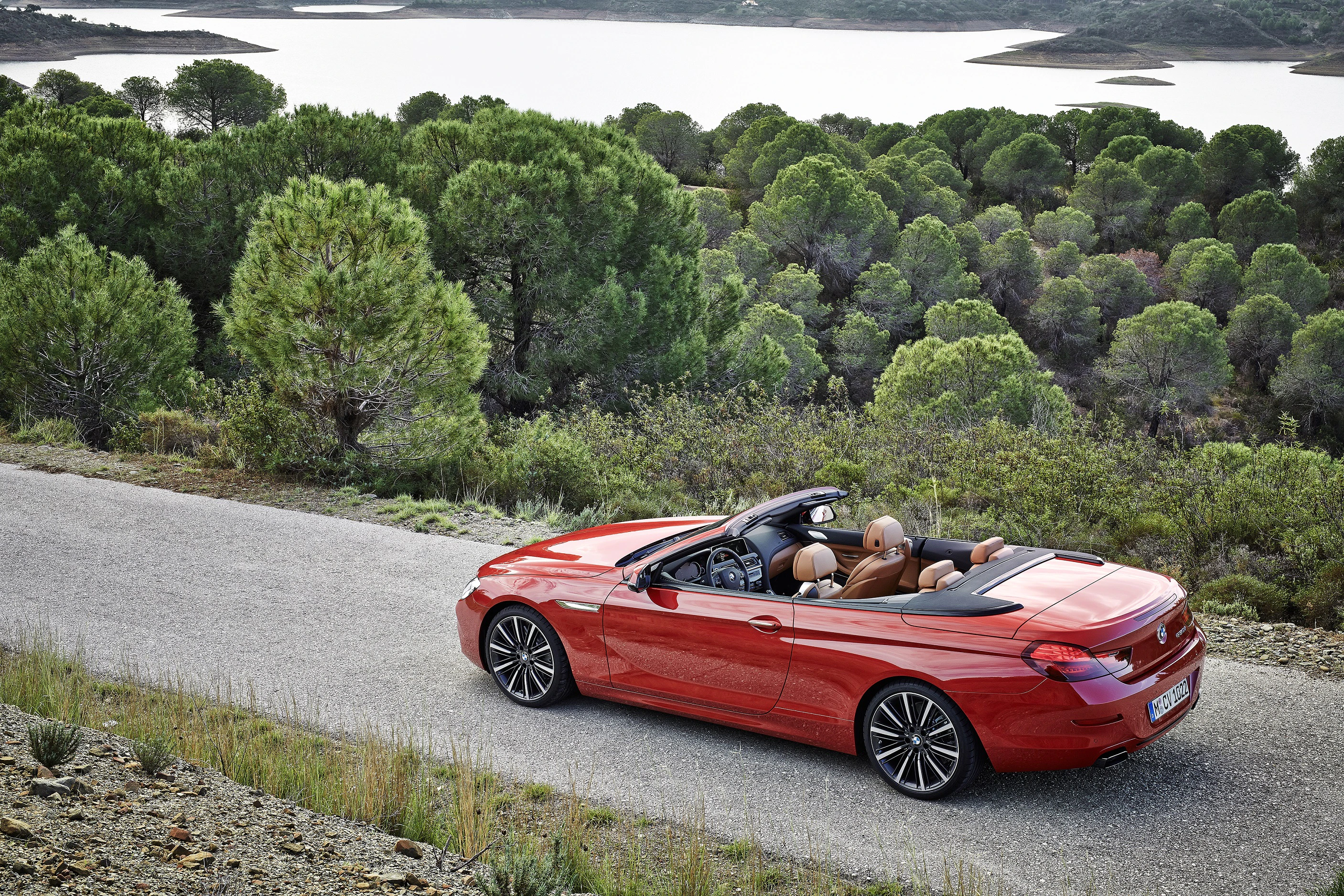 Roter Cabrio mit geöffnetem Verdeck fährt auf einer ländlichen Straße, umgeben von grünen Bäumen und einem See im Hintergrund.