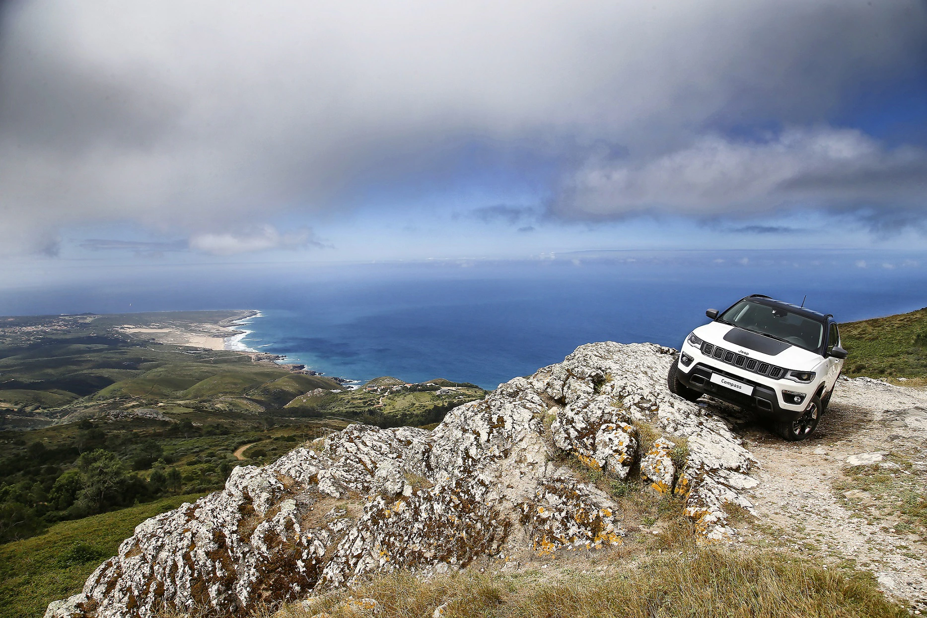 SUV auf felsigem Bergweg mit Meerblick und bewölktem Himmel im Hintergrund. Küstenlandschaft erstreckt sich in der Ferne.
