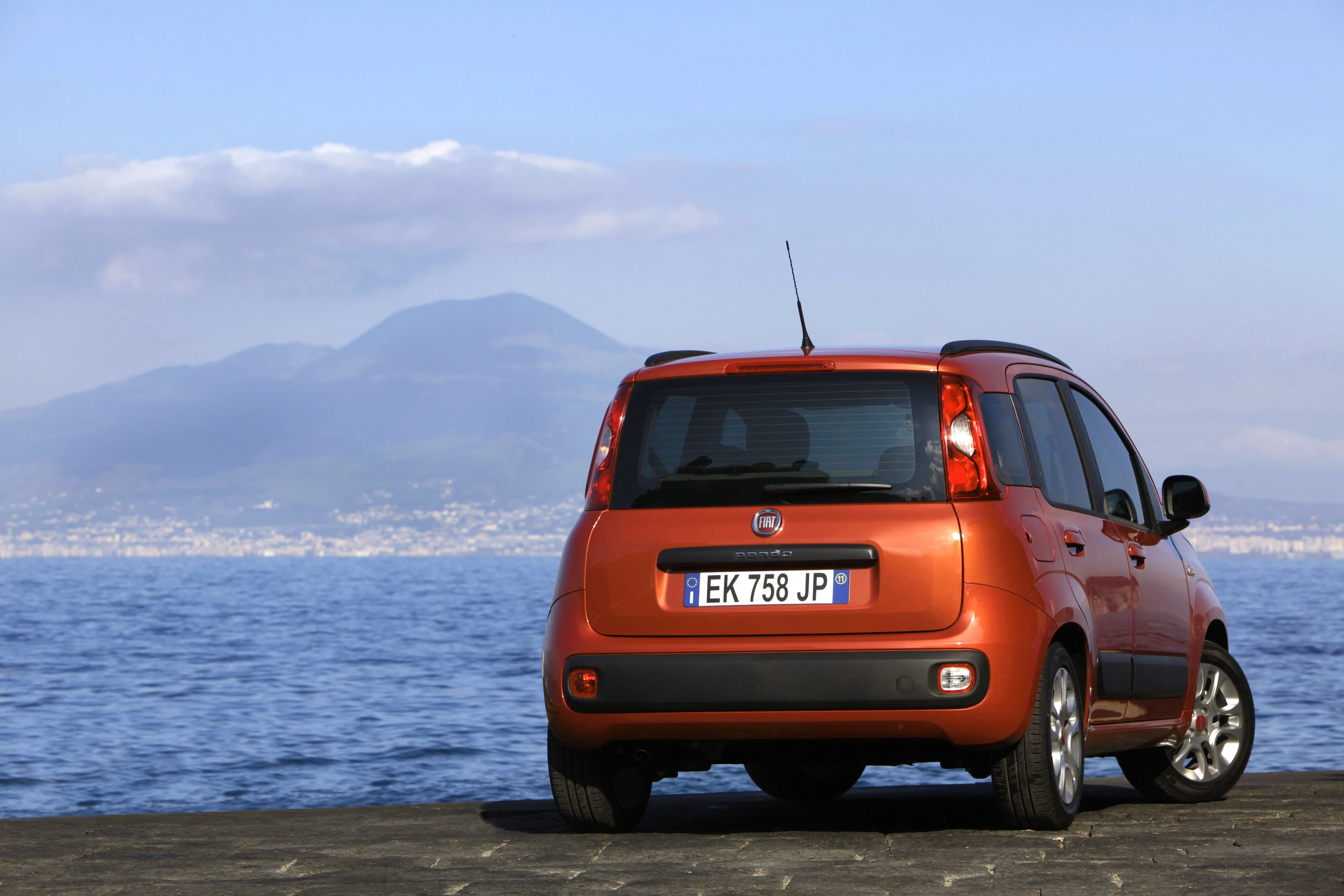 Ein orangefarbener Fiat parkt am Meer mit Blick auf einen Berg im Hintergrund.
