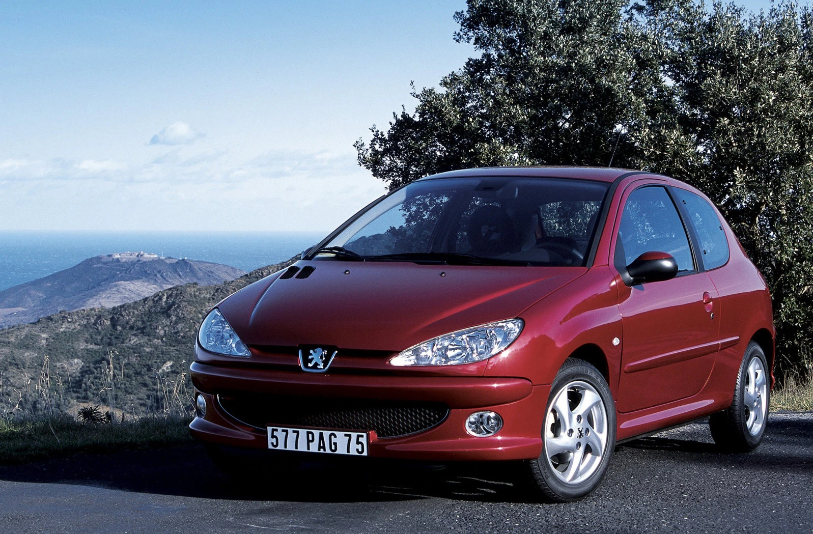 Roter Peugeot auf einer Bergstraße mit Meerblick. Im Hintergrund sind Hügel und Bäume unter blauem Himmel zu sehen.