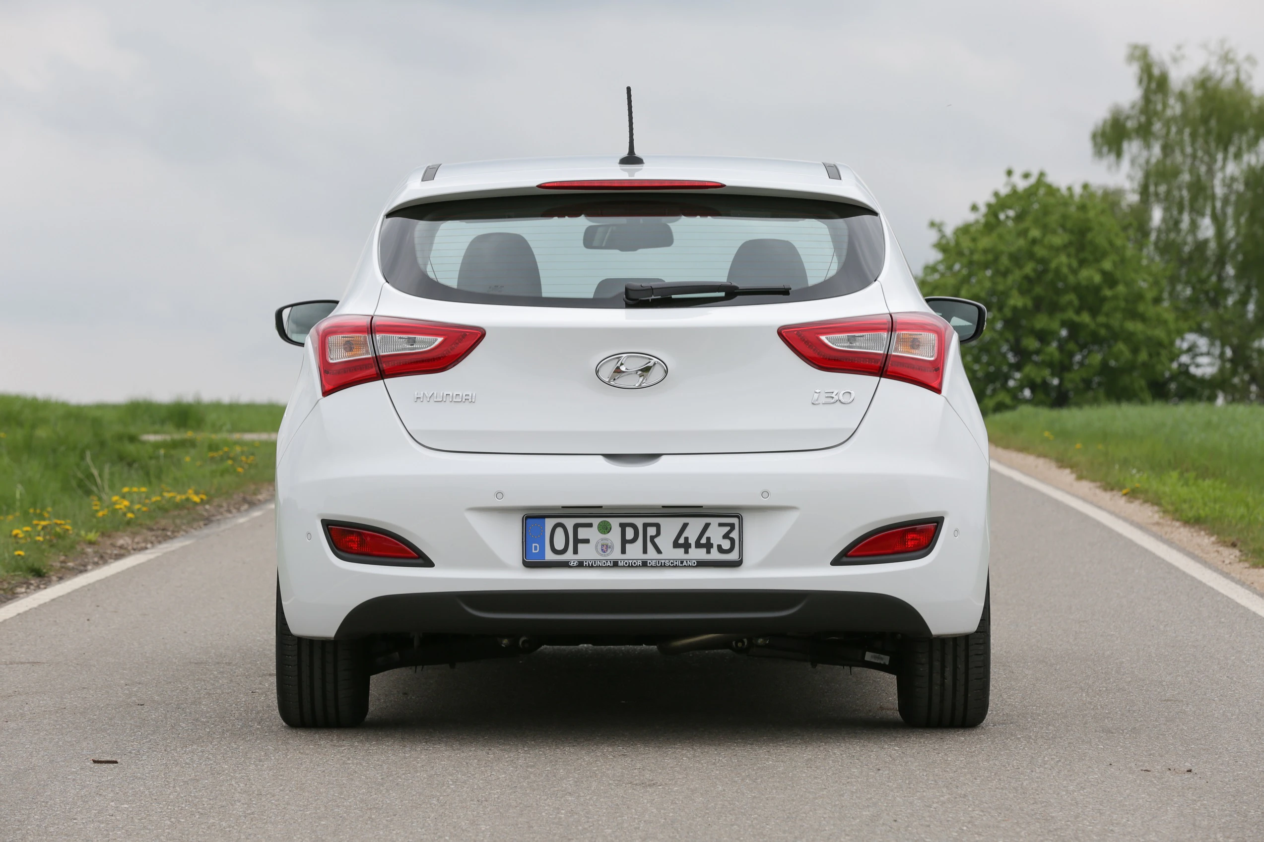 Rückansicht eines weißen Hyundai i30 auf einer Landstraße, Kennzeichen OF PR 443, umgeben von Gras und Bäumen unter bewölktem Himmel.