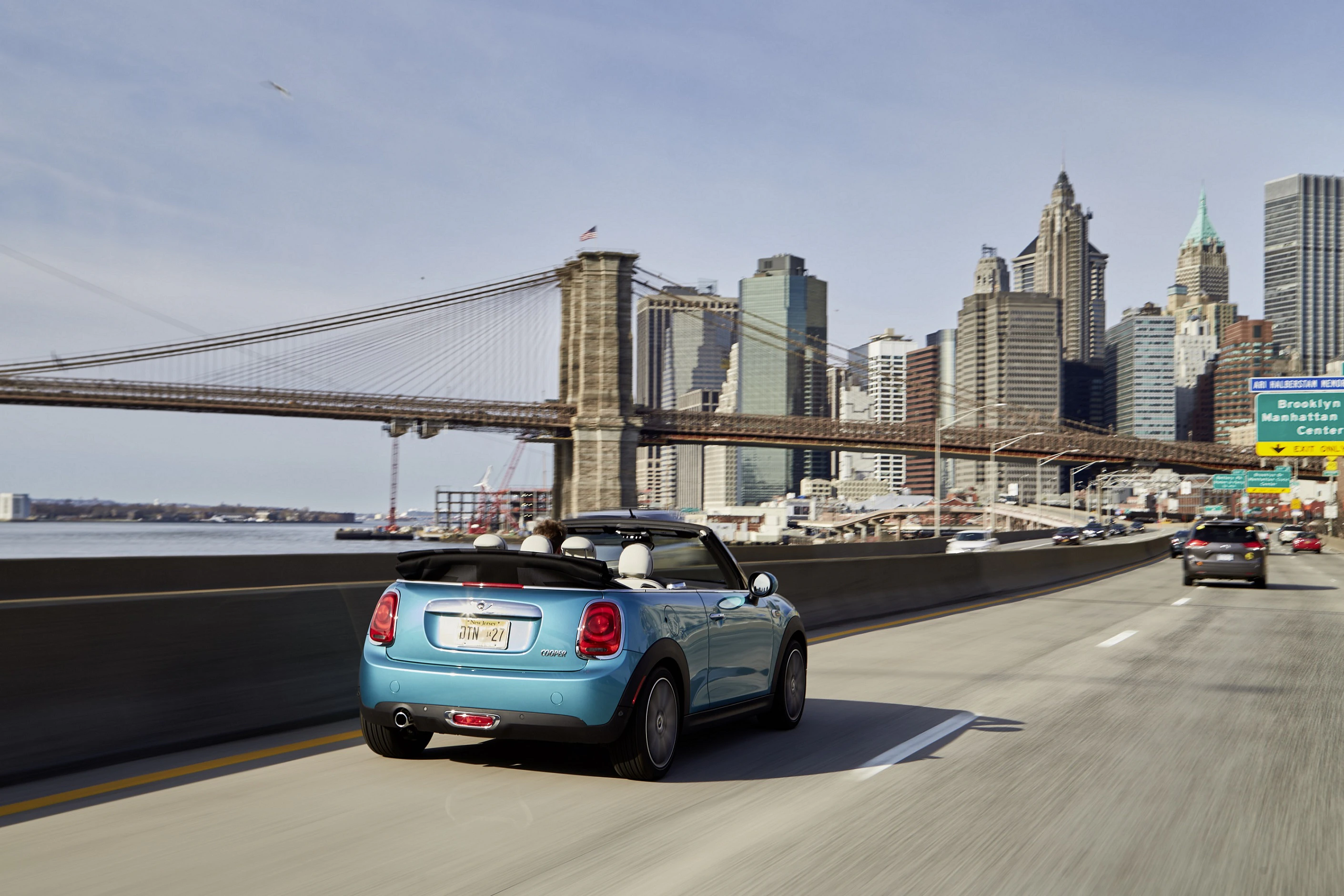 Blaues Cabrio fährt auf einer Stadtstraße in Richtung einer großen Brücke, mit der Skyline von New York im Hintergrund.