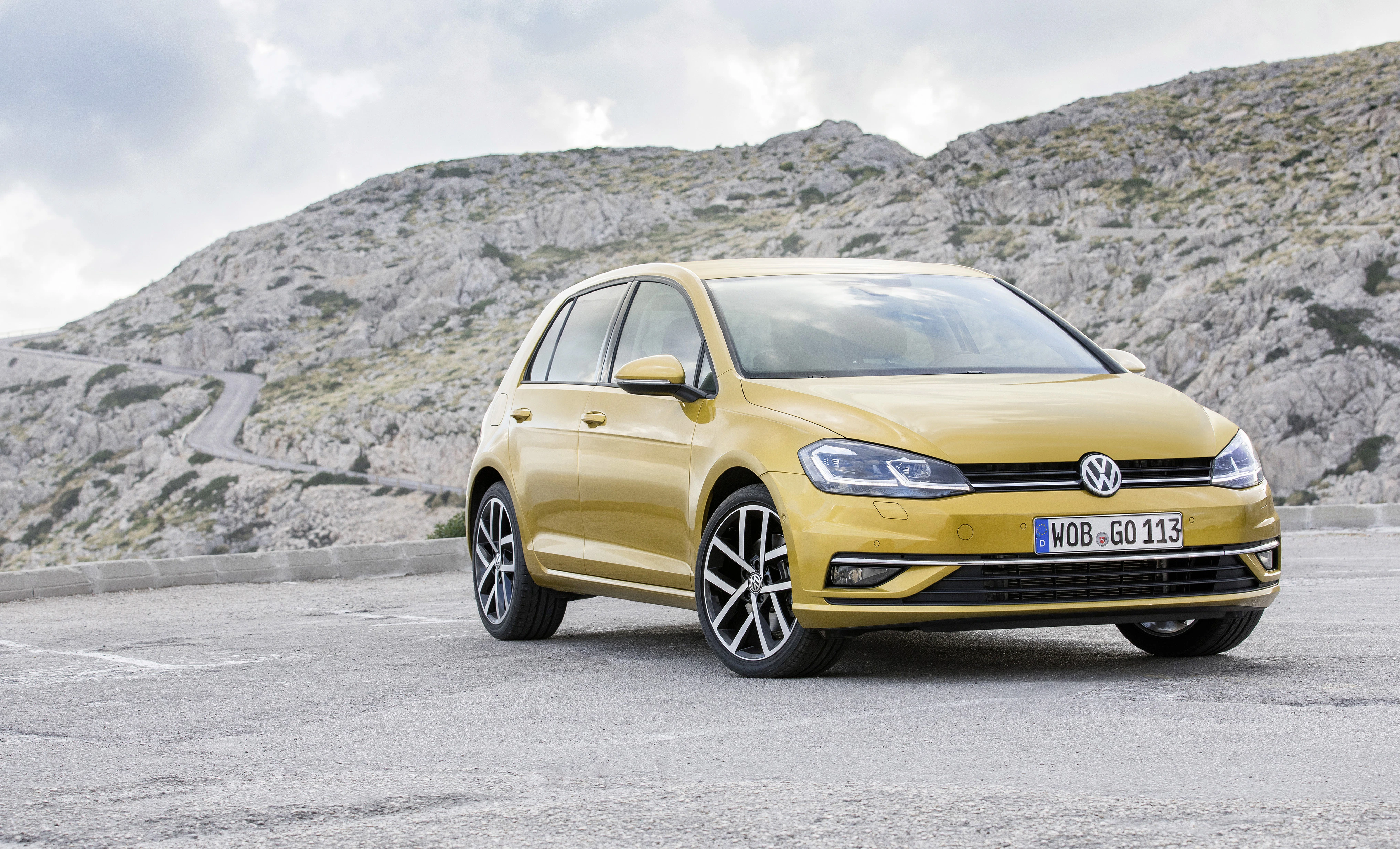 Gelber Volkswagen Golf steht auf einem Parkplatz vor einer felsigen Berglandschaft unter bewölktem Himmel.