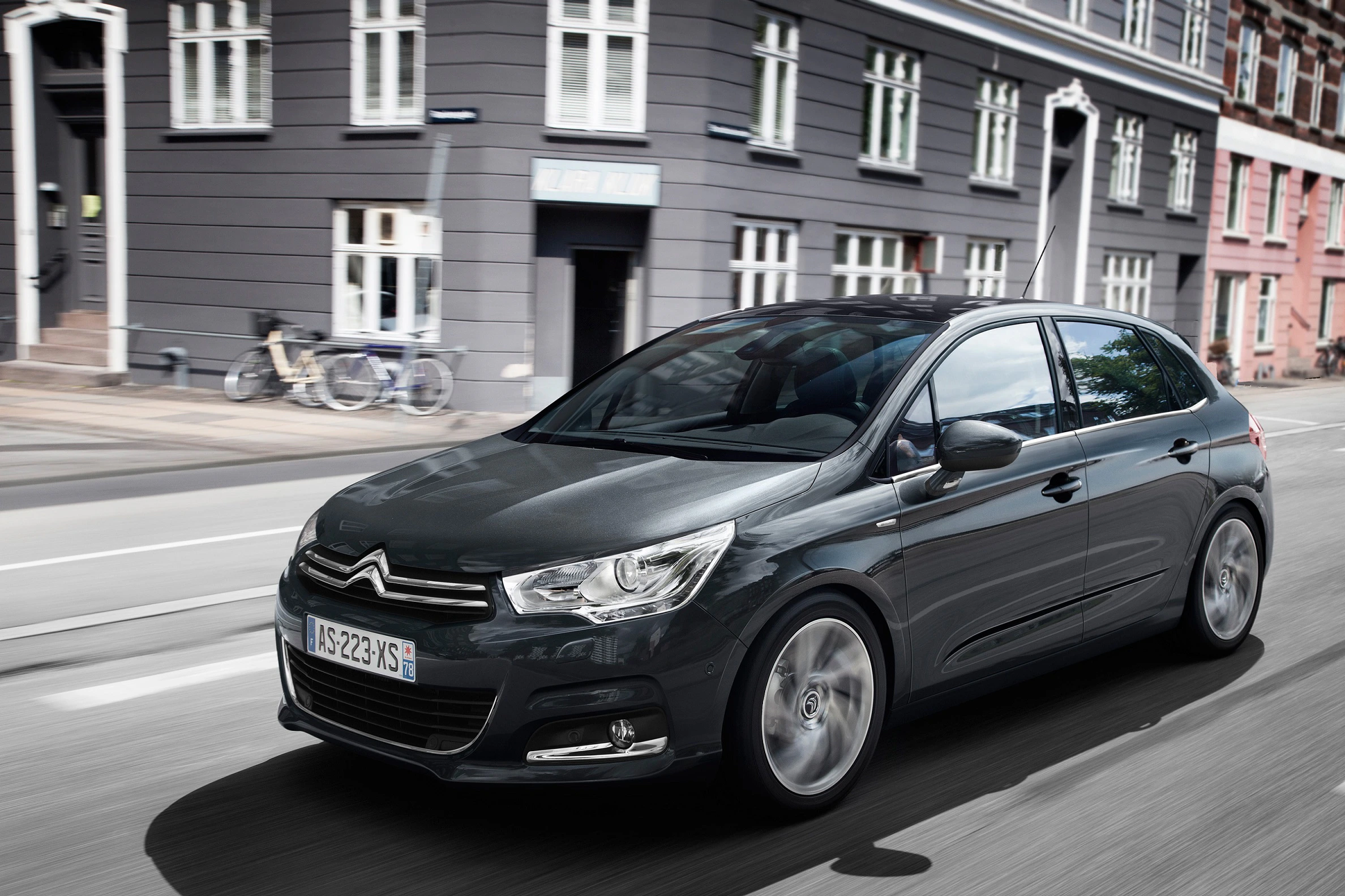 Ein dunkler Citroën C4 fährt auf einer städtischen Straße, vorbei an einem klassischen Gebäude mit Fahrrädern vor einer Fassade.