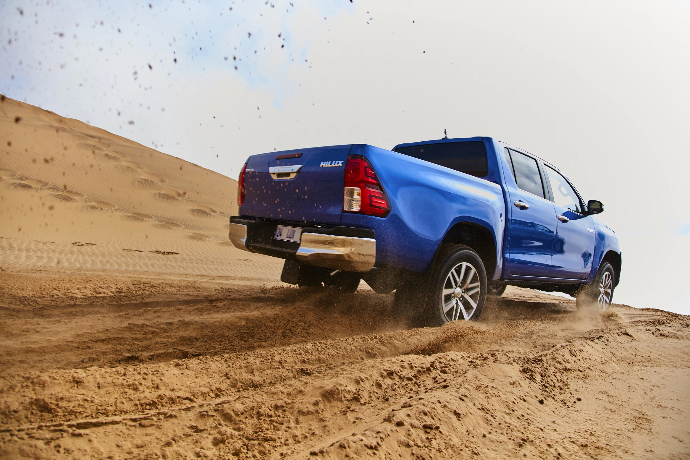 Blauer Pickup-Truck fährt durch sandige Wüste, wobei Sand aufgewirbelt wird. Fahrzeug in Bewegung auf geneigtem Gelände.
