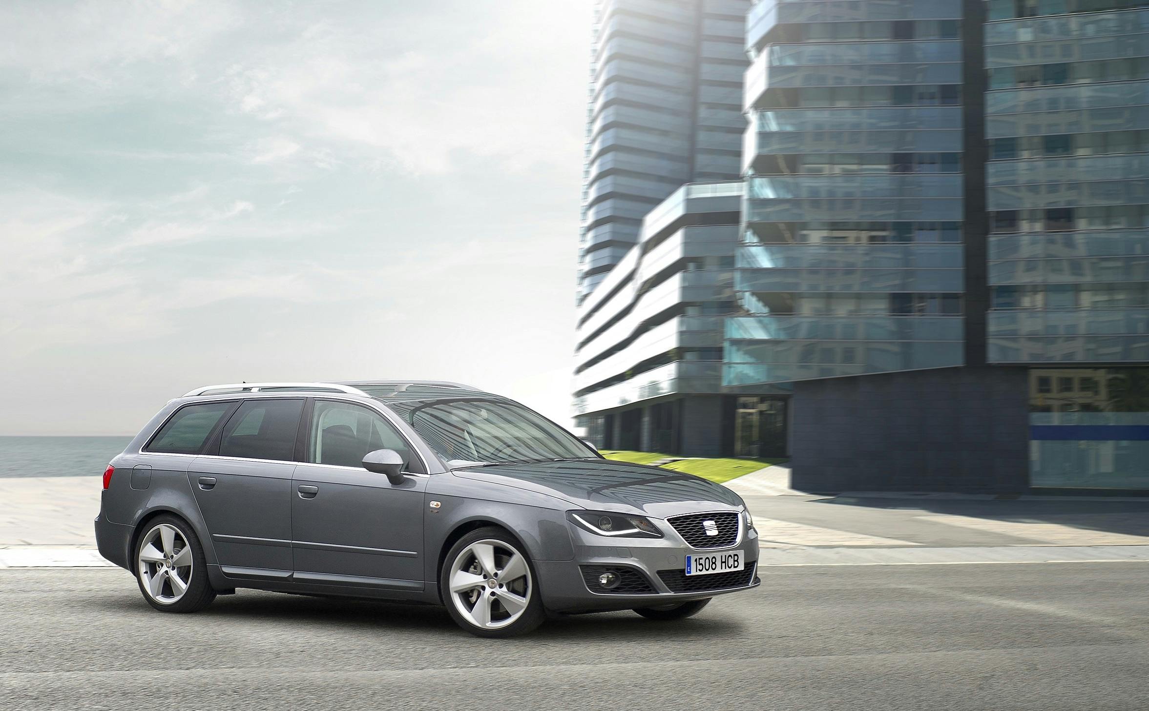 Grauer Kombi fährt auf städtischer Straße, moderne Glasgebäude im Hintergrund, sonniger Tag.