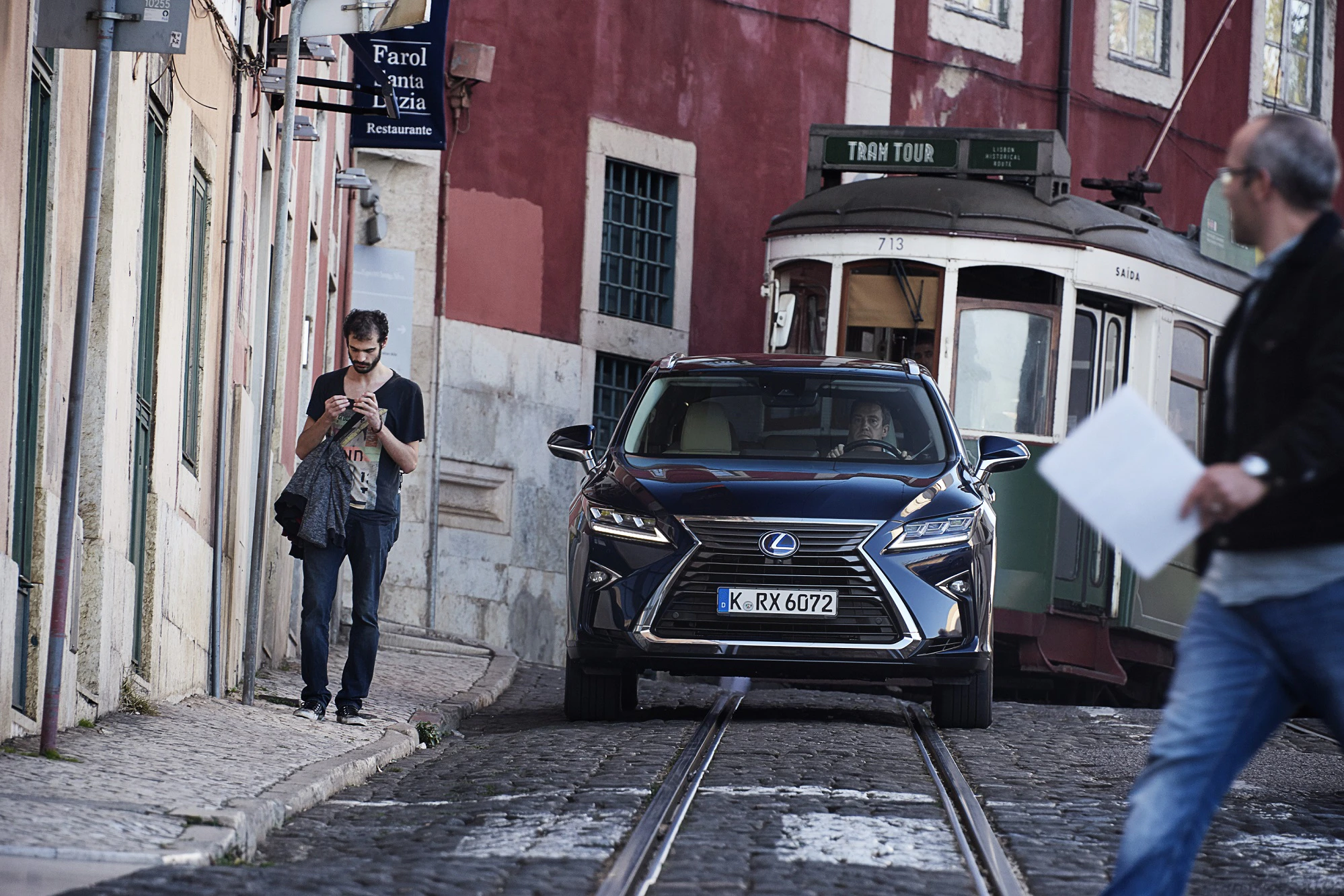 Ein Auto fährt eine enge Straße entlang, neben einem Fußgänger und einer vorbeifahrenden Straßenbahn.