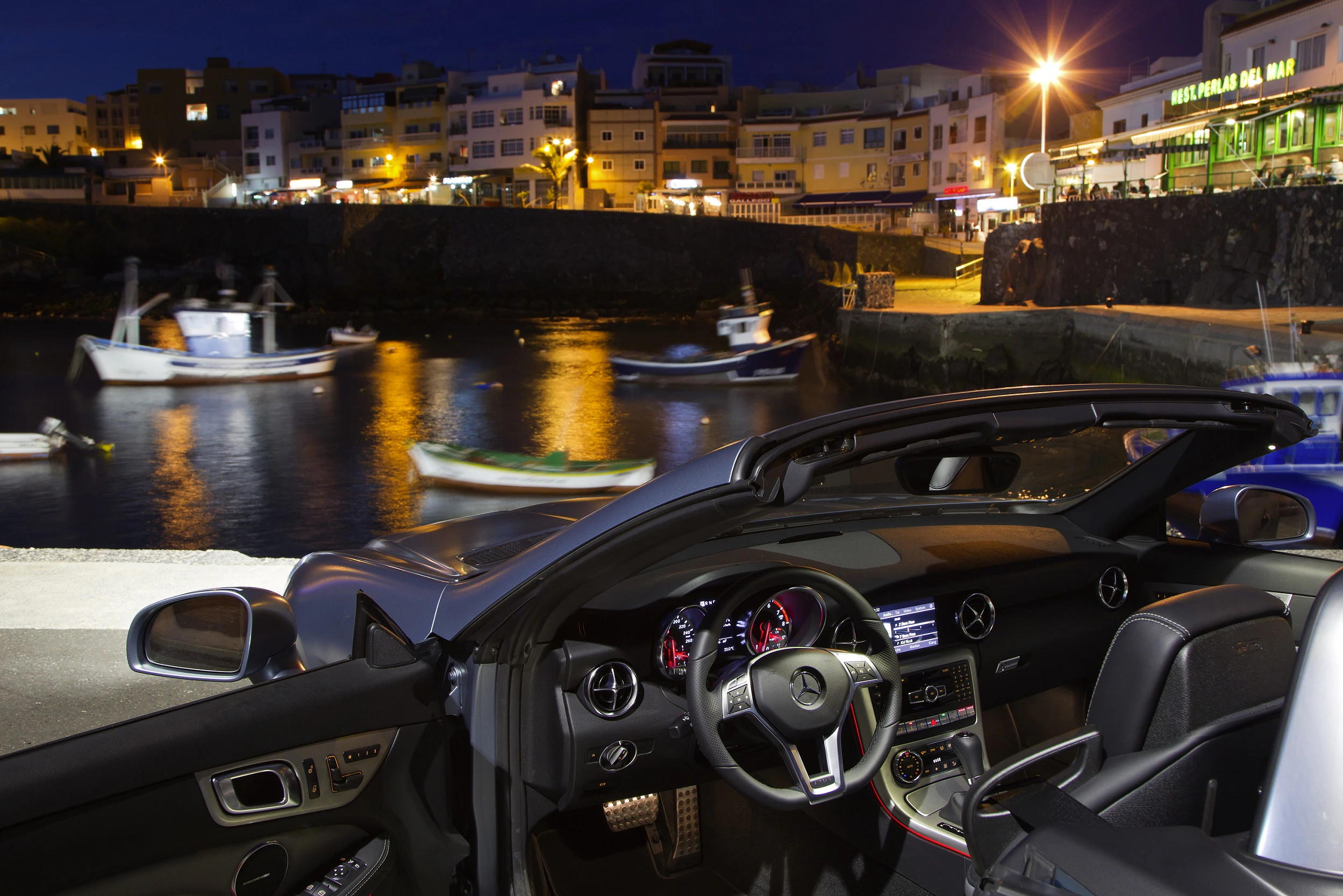 Ein Cabrio mit geöffnetem Verdeck steht am Wasser. Im Hintergrund sind beleuchtete Gebäude und Boote auf einem Fluss bei Nacht zu sehen.