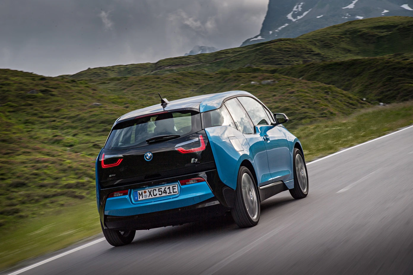 Blauer BMW i3 fährt auf einer malerischen Landstraße, umgeben von grünen Hügeln und bewölktem Himmel.