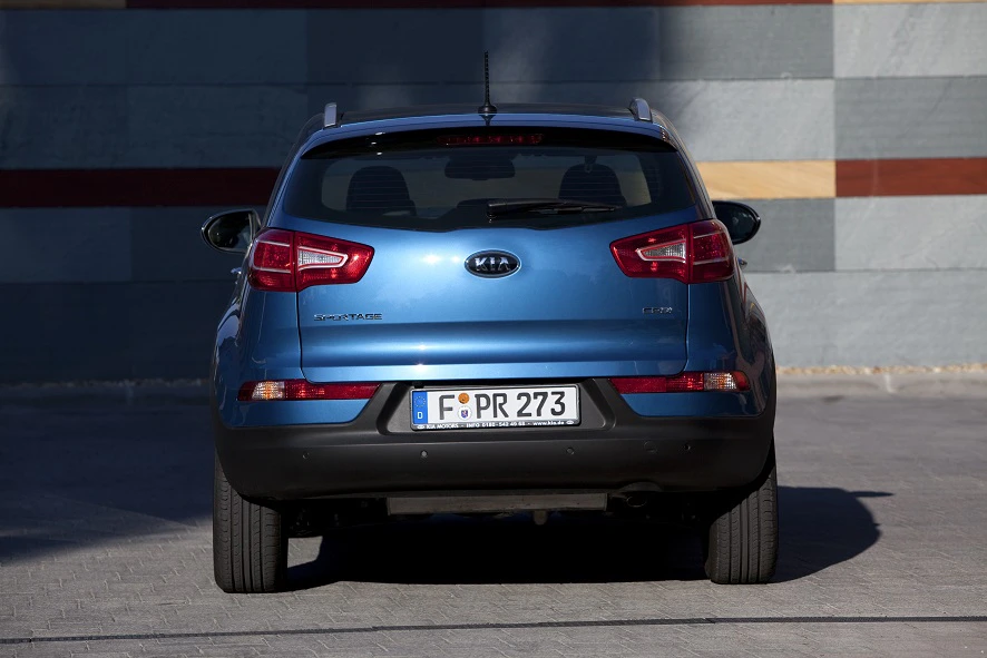 Blauer Kia Sportage von hinten, geparkt auf einem betonierten Platz. Bunt gemusterte Wand im Hintergrund.