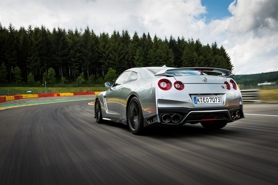 Silberner Sportwagen fährt schnell auf einer Rennstrecke mit Wald im Hintergrund und bunten Streckenmarkierungen.