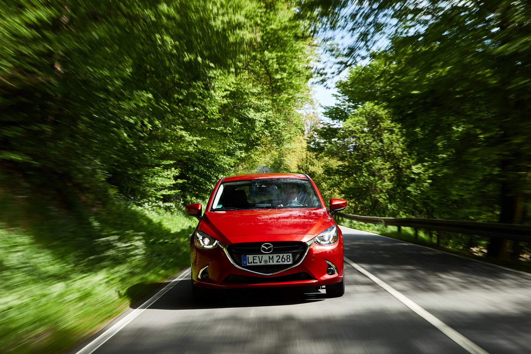 Roter Mazda fährt dynamisch auf einer von Bäumen gesäumten Straße. Geschwindigkeit und Natur vermitteln ein Gefühl von Freiheit.