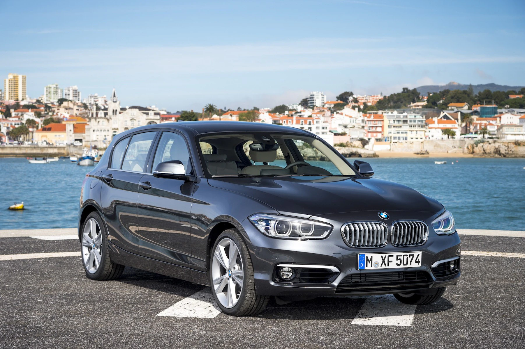 Grauer BMW 1er auf Parkplatz am Wasser, im Hintergrund eine Stadt mit weißen Gebäuden und blauem Himmel.
