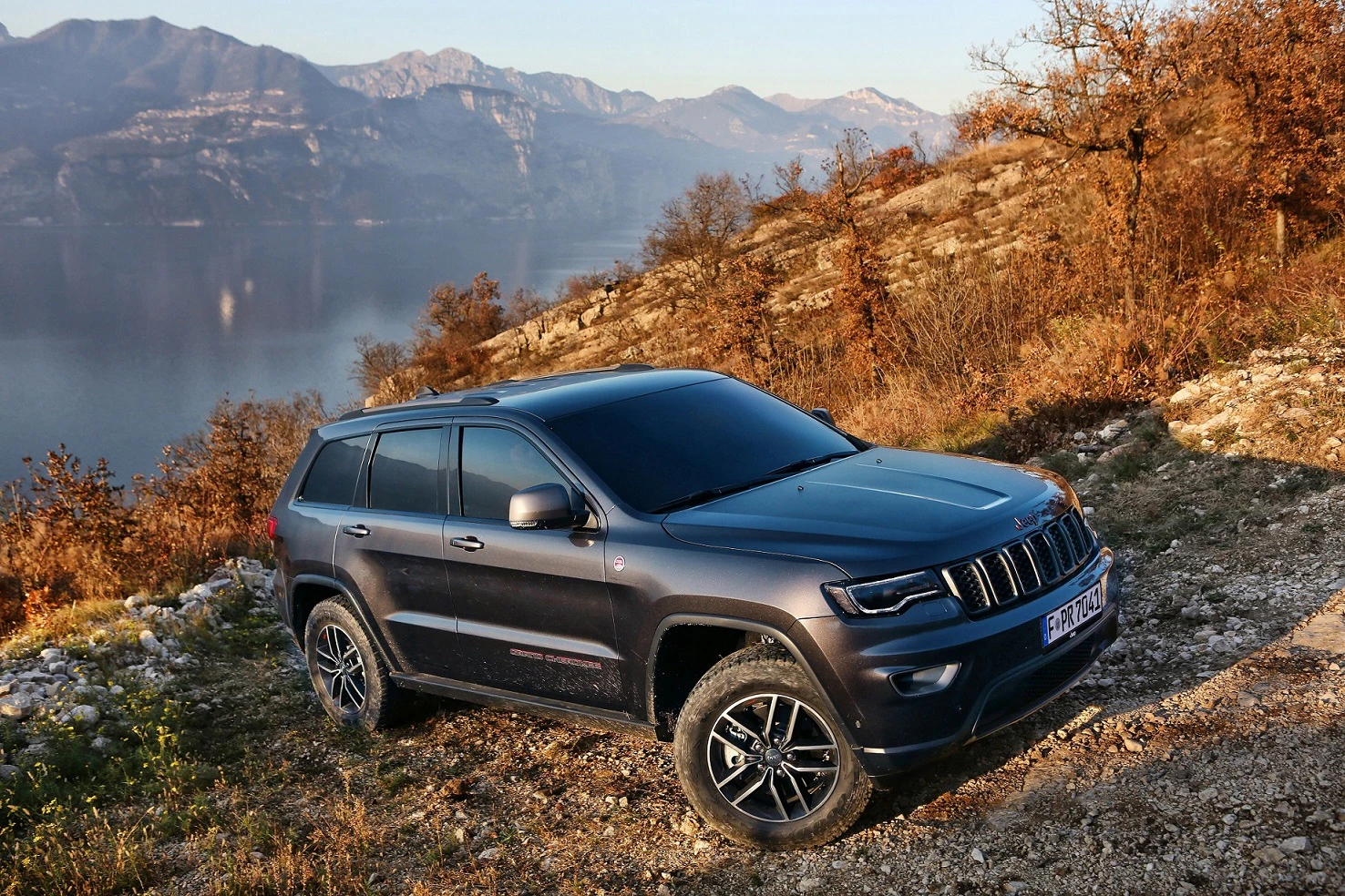 Dunkelblauer SUV parkt im Herbst auf einem steinigen Hügel mit Blick auf einen ruhigen See und Berge im Hintergrund.