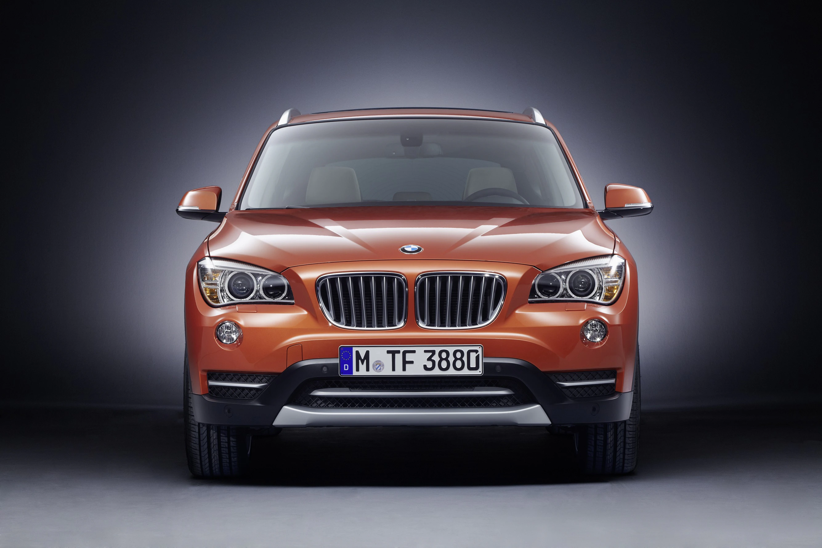 Orangefarbener BMW SUV, frontale Ansicht, auf dunklem Hintergrund. Fahrzeugbeleuchtung ist eingeschaltet.