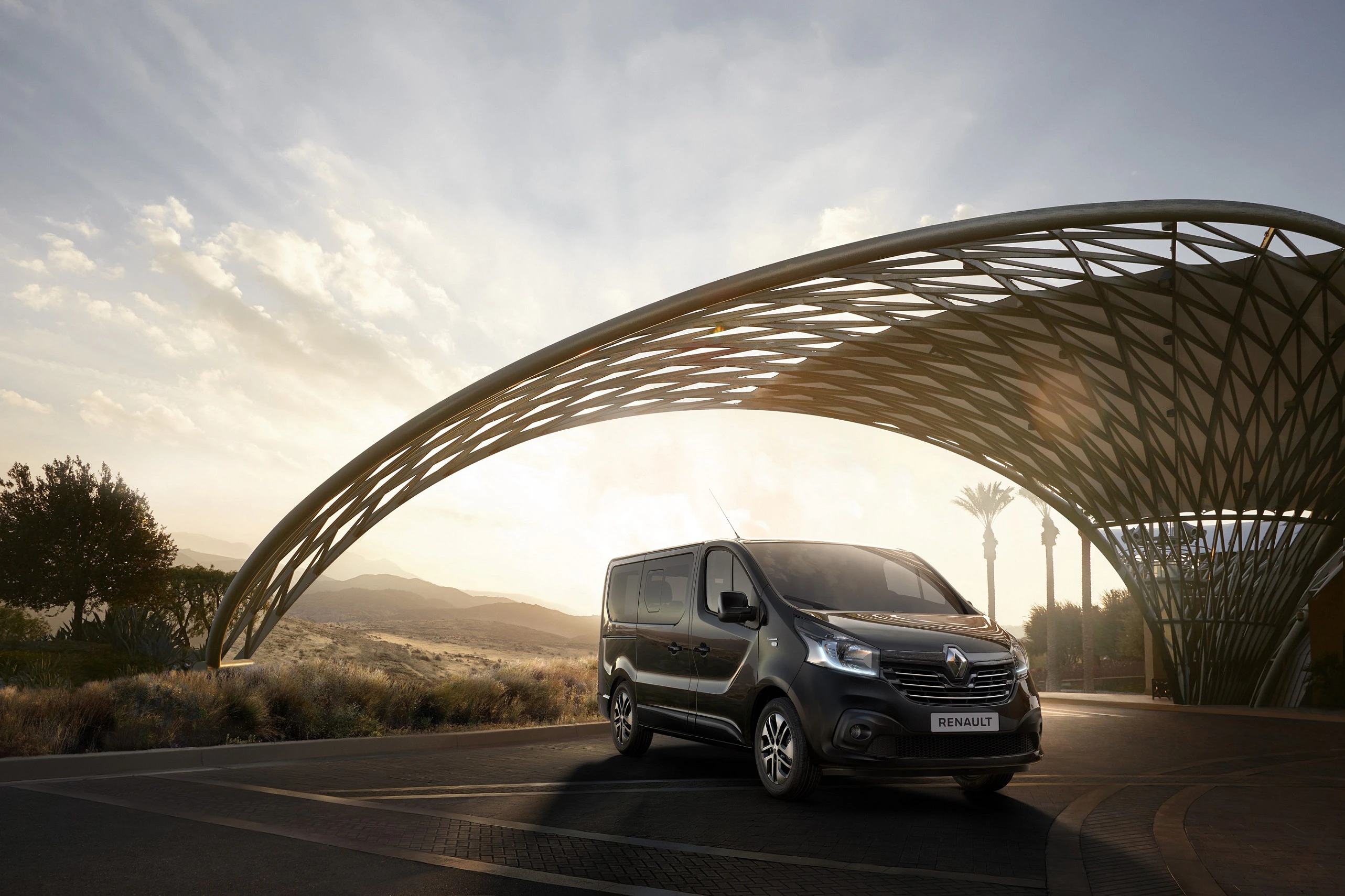 Ein schwarzer Renault-Transporter fährt bei Sonnenuntergang unter einer futuristischen, geschwungenen Struktur entlang einer Straße.
