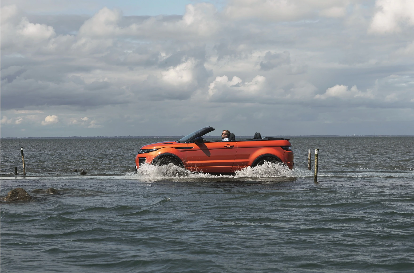 Orangefarbenes Cabrio fährt durch flaches Wasser auf einer überfluteten Straße unter wolkigem Himmel.