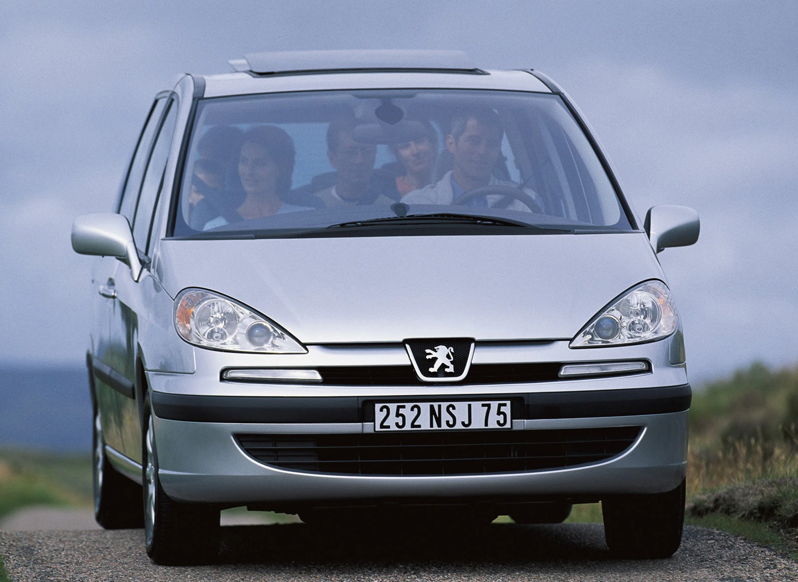 Silberner Peugeot Van fährt auf einer Landstraße. Mehrere Personen sind im Fahrzeug. Überwiegend bewölkter Himmel im Hintergrund.