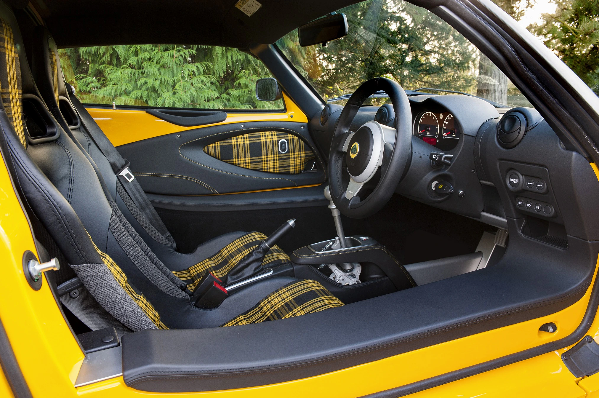 Gelber Sportwagen-Innenraum mit schwarzem und kariertem Stoff. Linkslenker, Schalthebel und moderne Anzeigen. Hintergrund mit Bäumen.