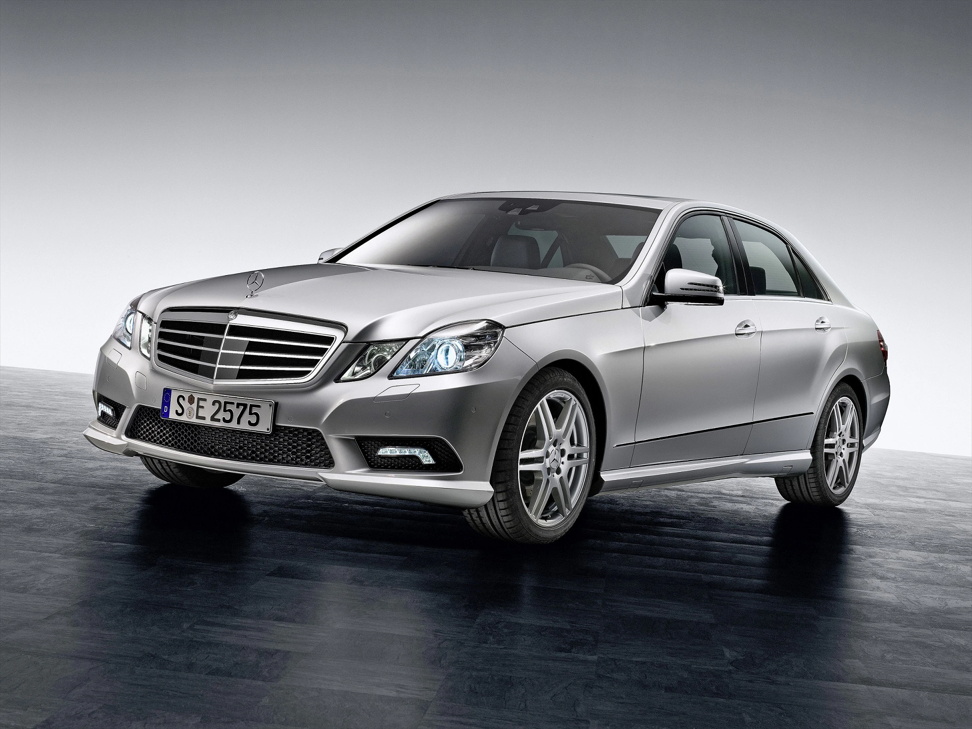 Silberner Mercedes-Benz Limousine auf grauem Hintergrund, frontale Ansicht. Fahrzeug hat markante Scheinwerfer und stilvolle Alufelgen.