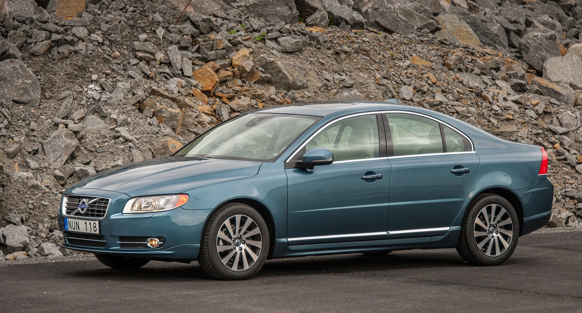 Blauer Volvo S80, Limousine, geparkt auf Asphalt vor einer felsigen, grauen Hintergrundszenerie.