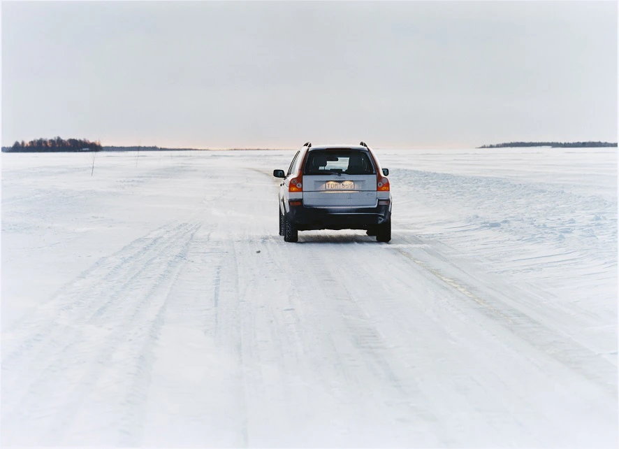 Ein Auto fährt auf einer schneebedeckten, vereisten Straße in einer weiten, leeren Landschaft. Der Himmel ist bewölkt und grau.