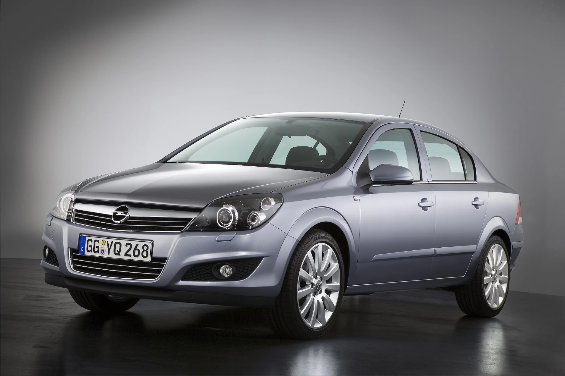 Silberner Opel Astra Limousine in einem Studio mit eingeschalteten Scheinwerfern. Vorder- und Seitenansicht auf dunklem Boden.