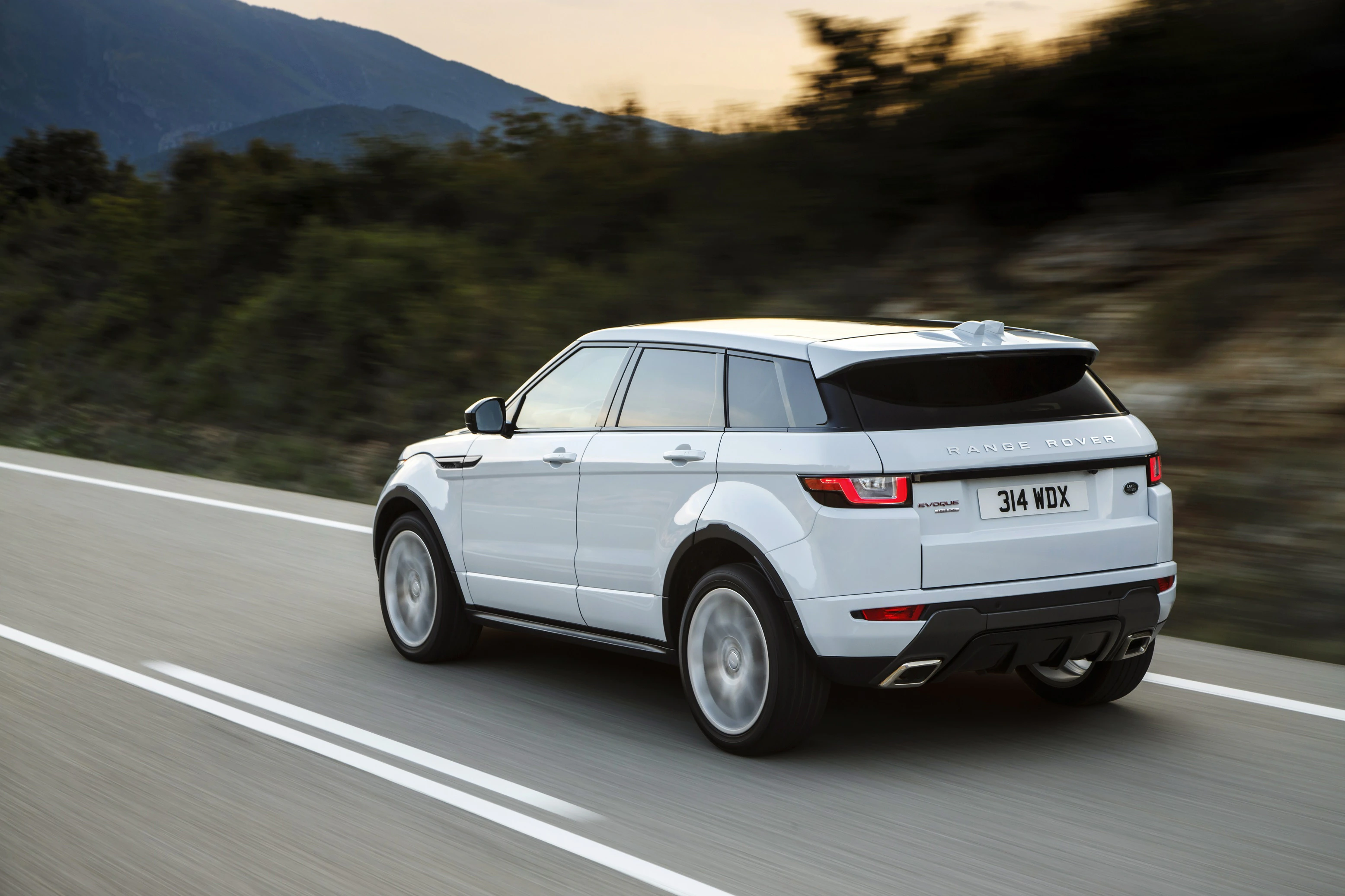 Weißer SUV fährt auf einer Landstraße bei Tageslicht, mit Bäumen und Hügeln im Hintergrund.