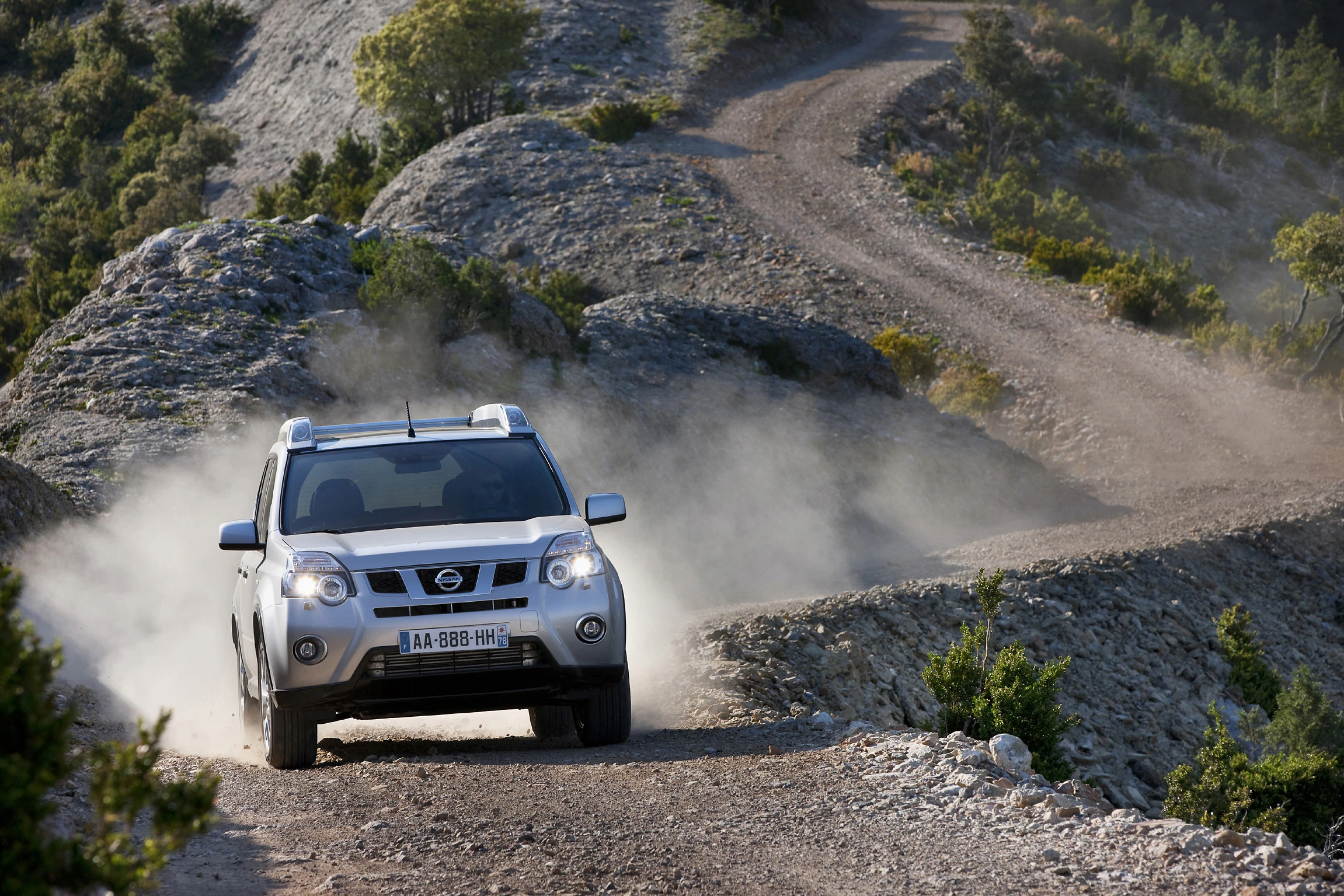Ein silberner SUV fährt auf einer staubigen, kurvigen Bergstraße durch eine felsige und bewaldete Landschaft.