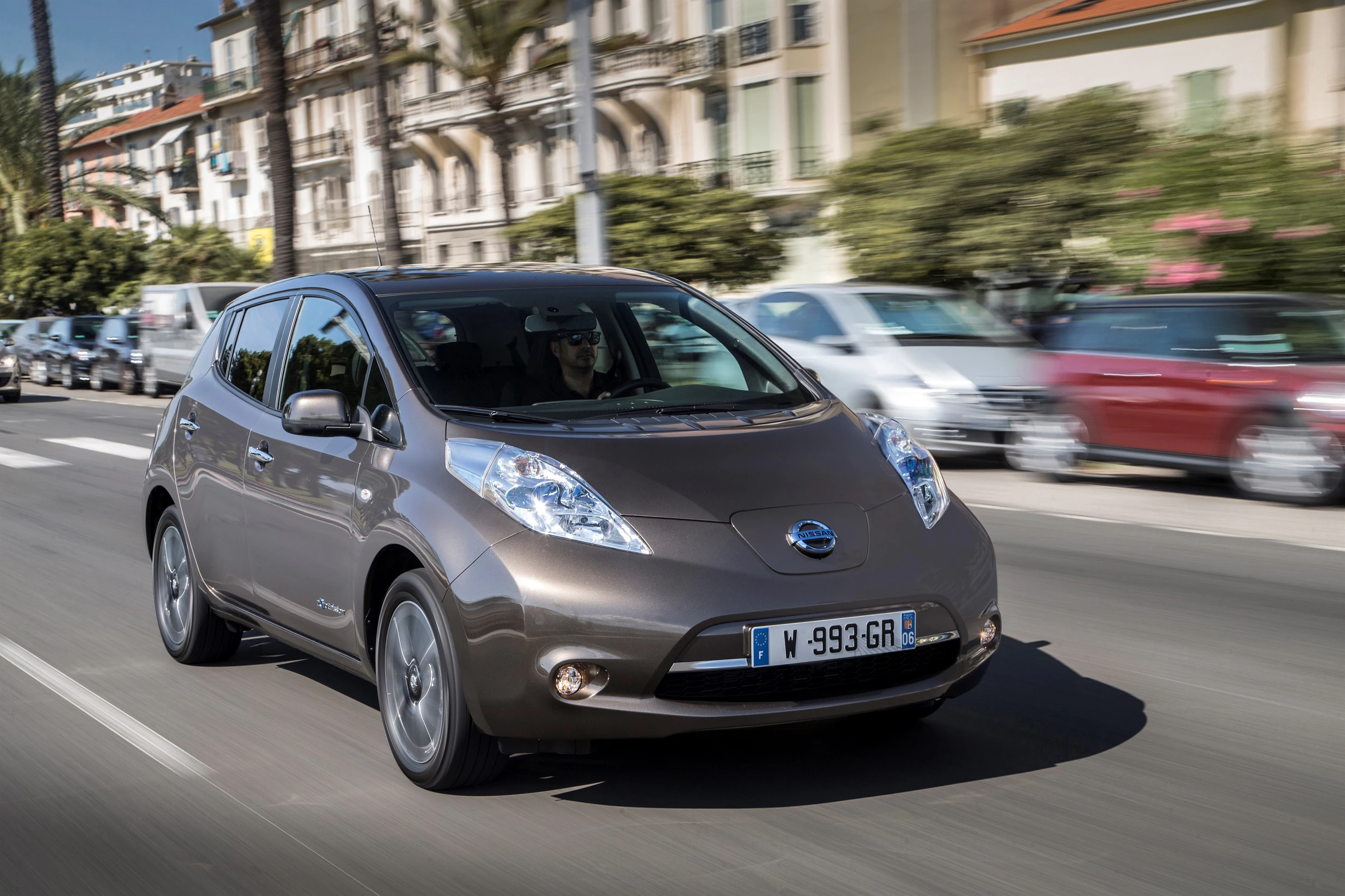 Brauner Nissan Leaf fährt auf städtischer Straße, umgeben von Autos und Gebäuden im Hintergrund. Sonniges Wetter, Fahrer sichtbar.