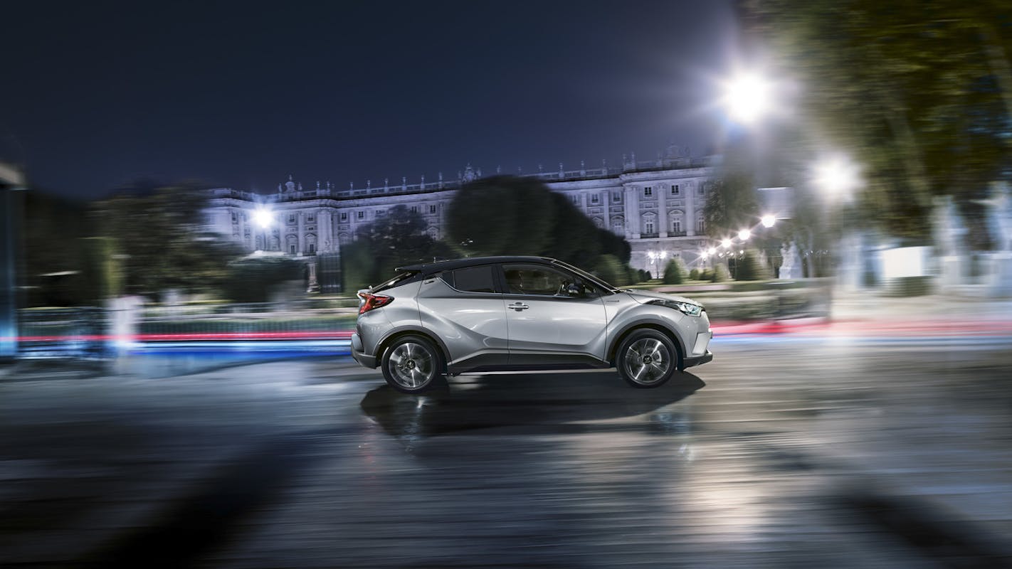 Silbernes Auto fährt nachts durch eine beleuchtete Stadtstraße, verschwommener Hintergrund mit historischem Gebäude.