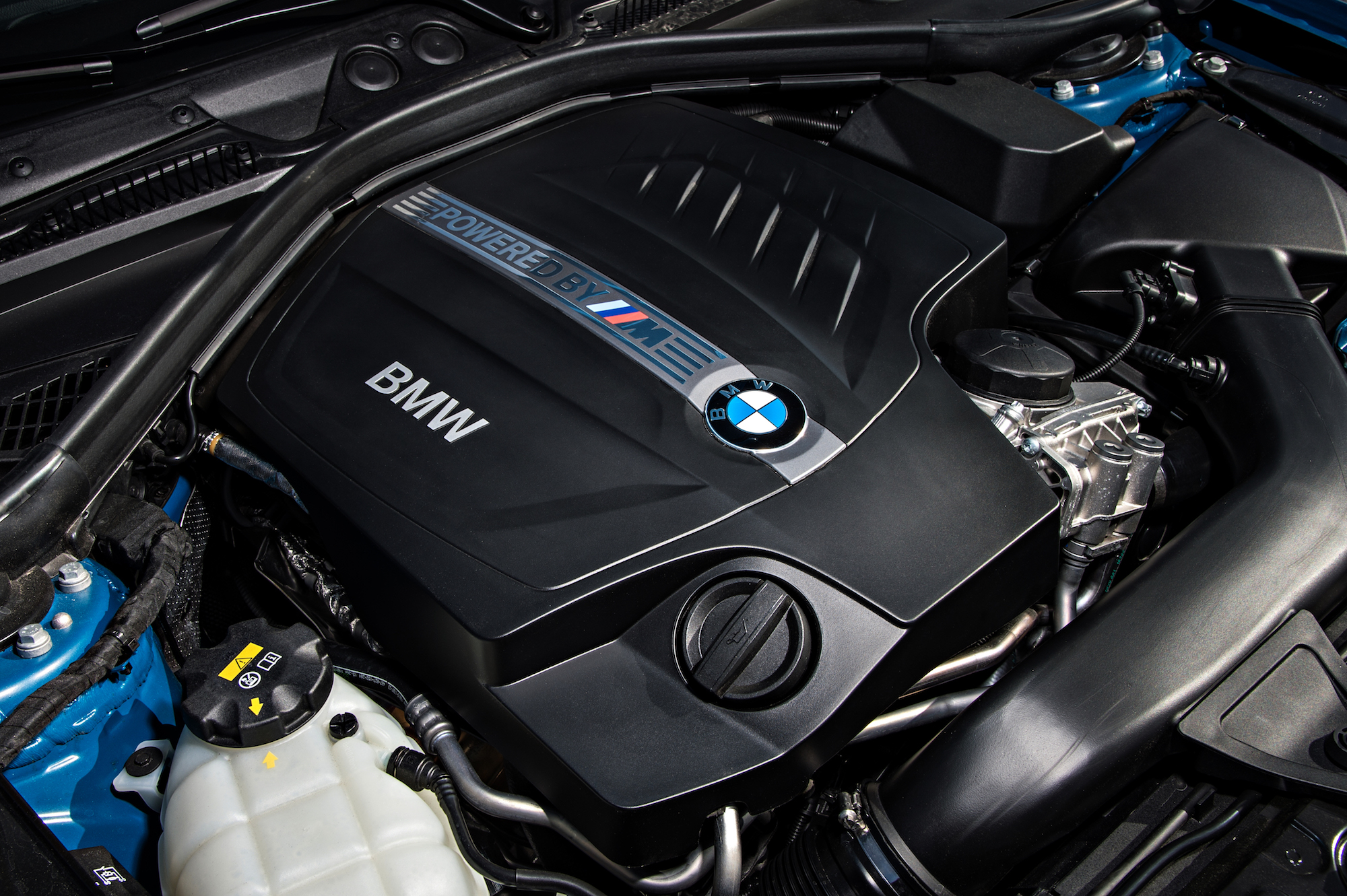BMW-Motorraum mit schwarzer Abdeckung, BMW-Logo und "POWERED BY M" Schriftzug, umgeben von blauen und schwarzen Bauteilen.