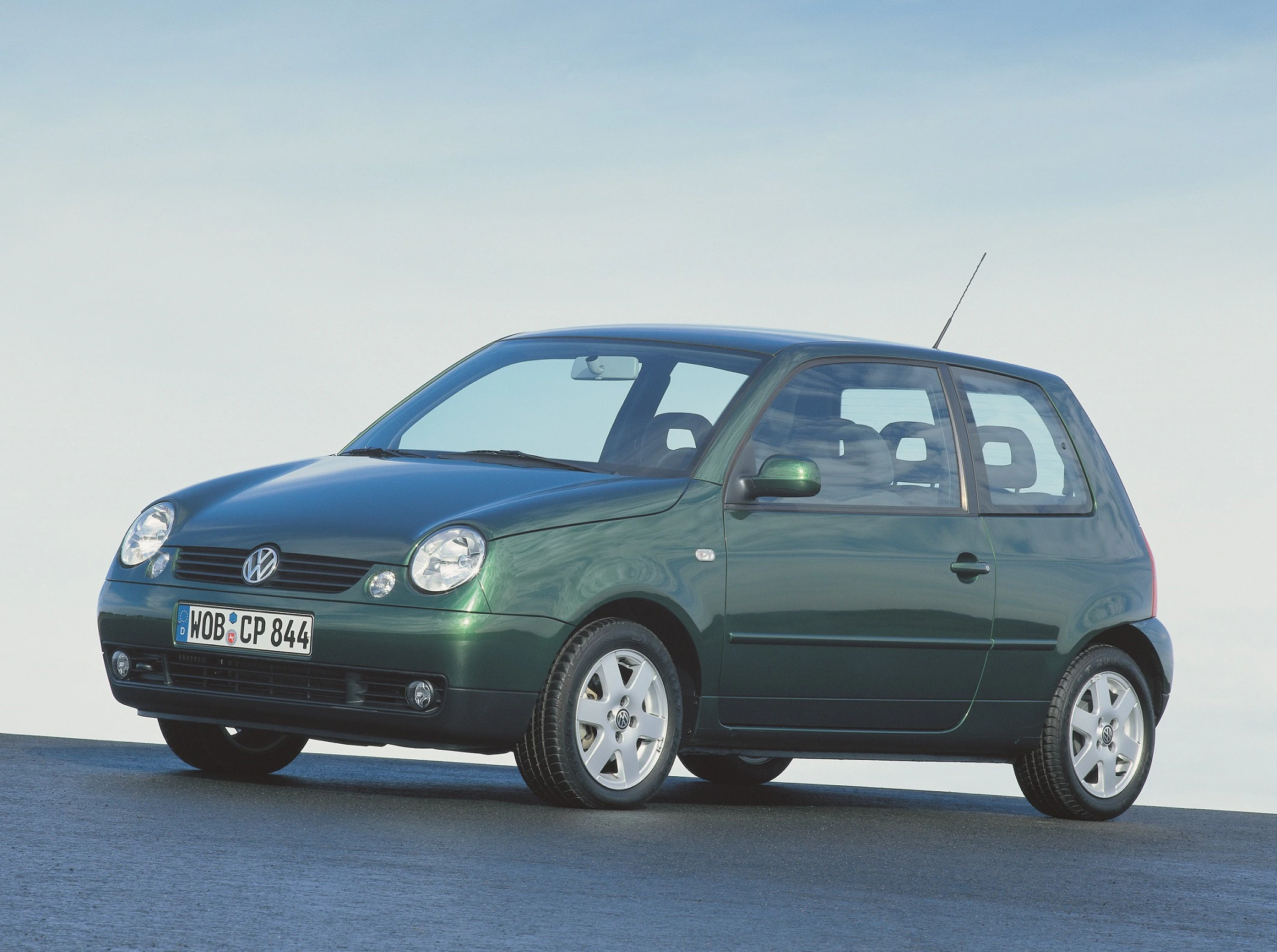 Grüner VW Lupo, Seitenansicht. Parkt auf einer asphaltierten Fläche unter blauem Himmel. Fahrzeug hat drei Türen.