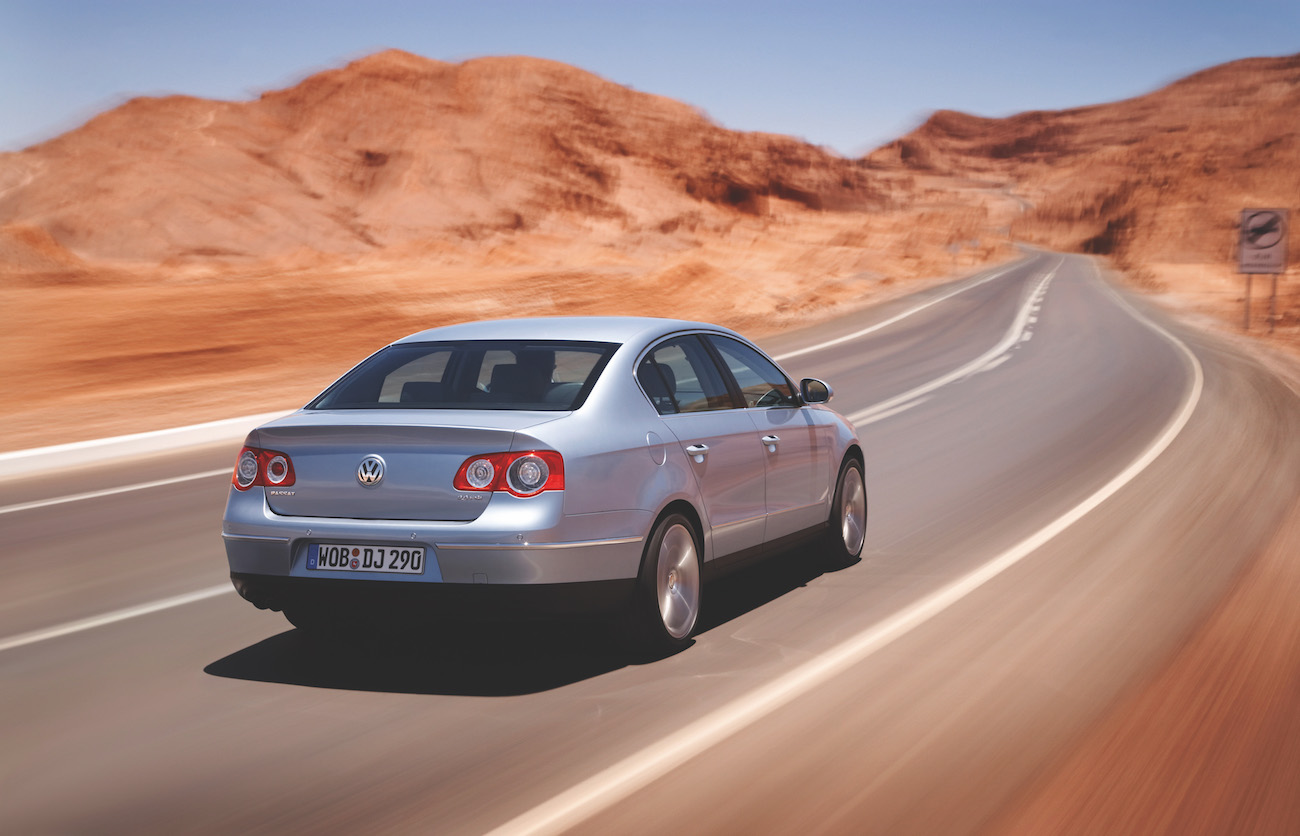 Silberner Volkswagen fährt auf einer kurvigen, einsamen Wüstenstraße unter klarem Himmel.