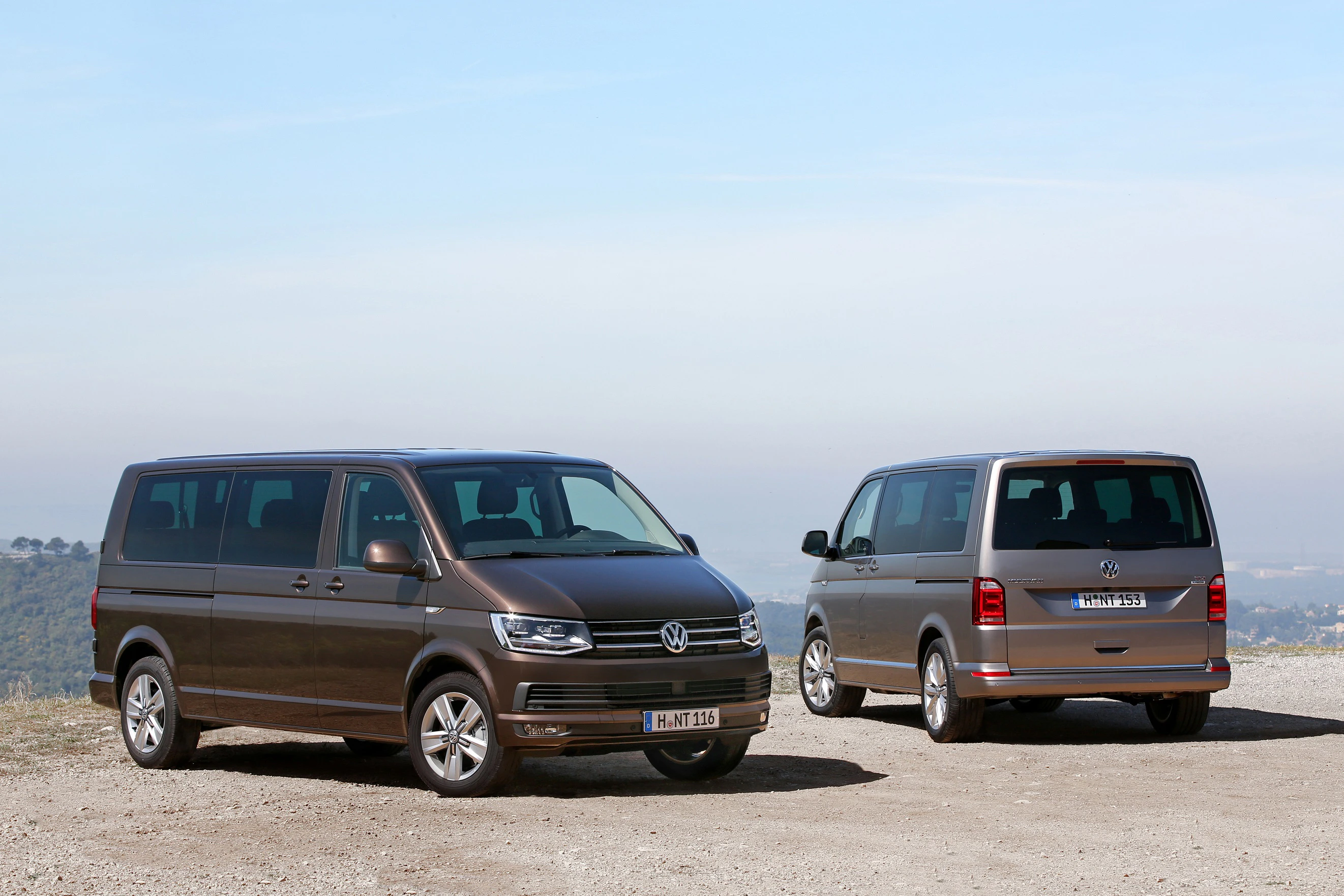 Zwei braune VW-Transporter stehen auf einem Hügel mit Fernblick. Einer ist zur Seite gedreht, der andere zeigt die Rückseite.