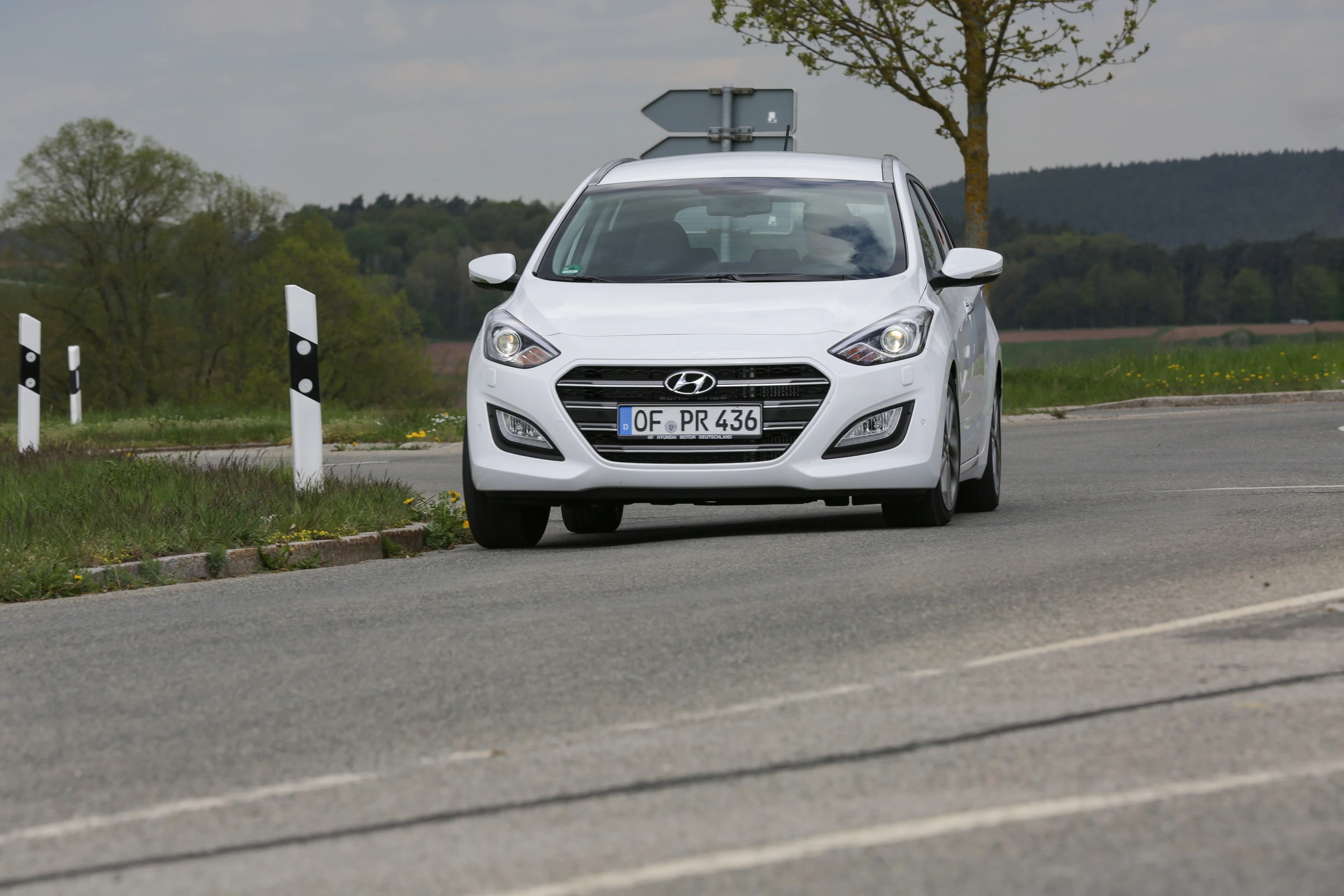 Ein weißer Hyundai fährt auf einer Landstraße. Im Hintergrund sind Bäume und ein Schild.