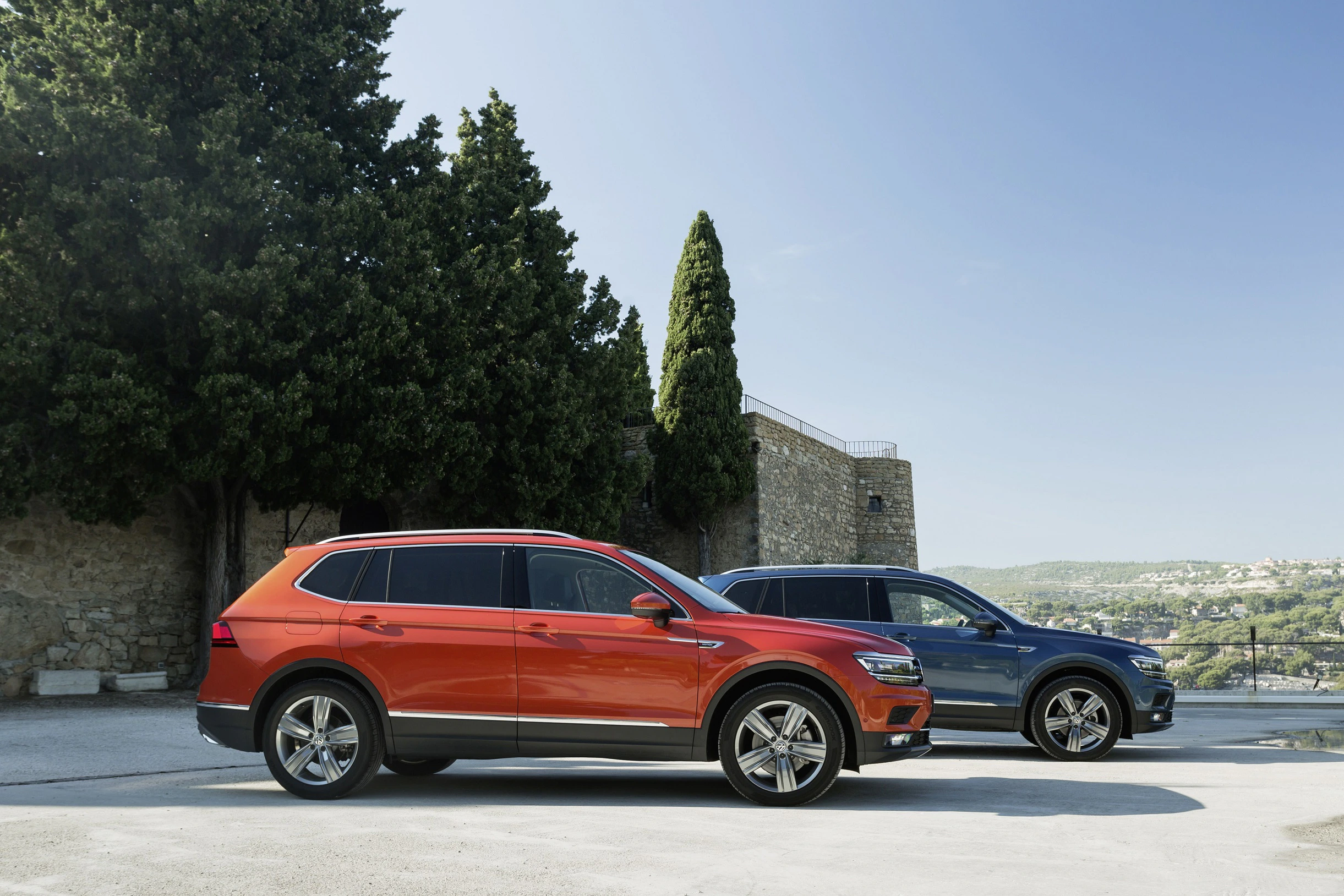 Zwei SUV-PKW, einer rot und einer blau, stehen nebeneinander vor einer Steinmauer und hohen Bäumen mit klarem Himmel im Hintergrund.
