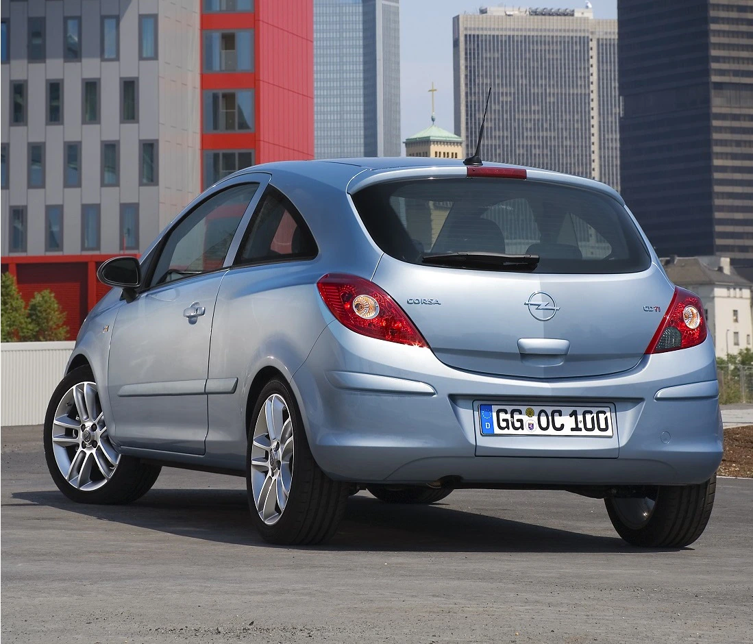 Silberner Opel Corsa parkt auf einem städtischen Parkplatz, umgeben von modernen Gebäuden und Wolkenkratzern im Hintergrund.