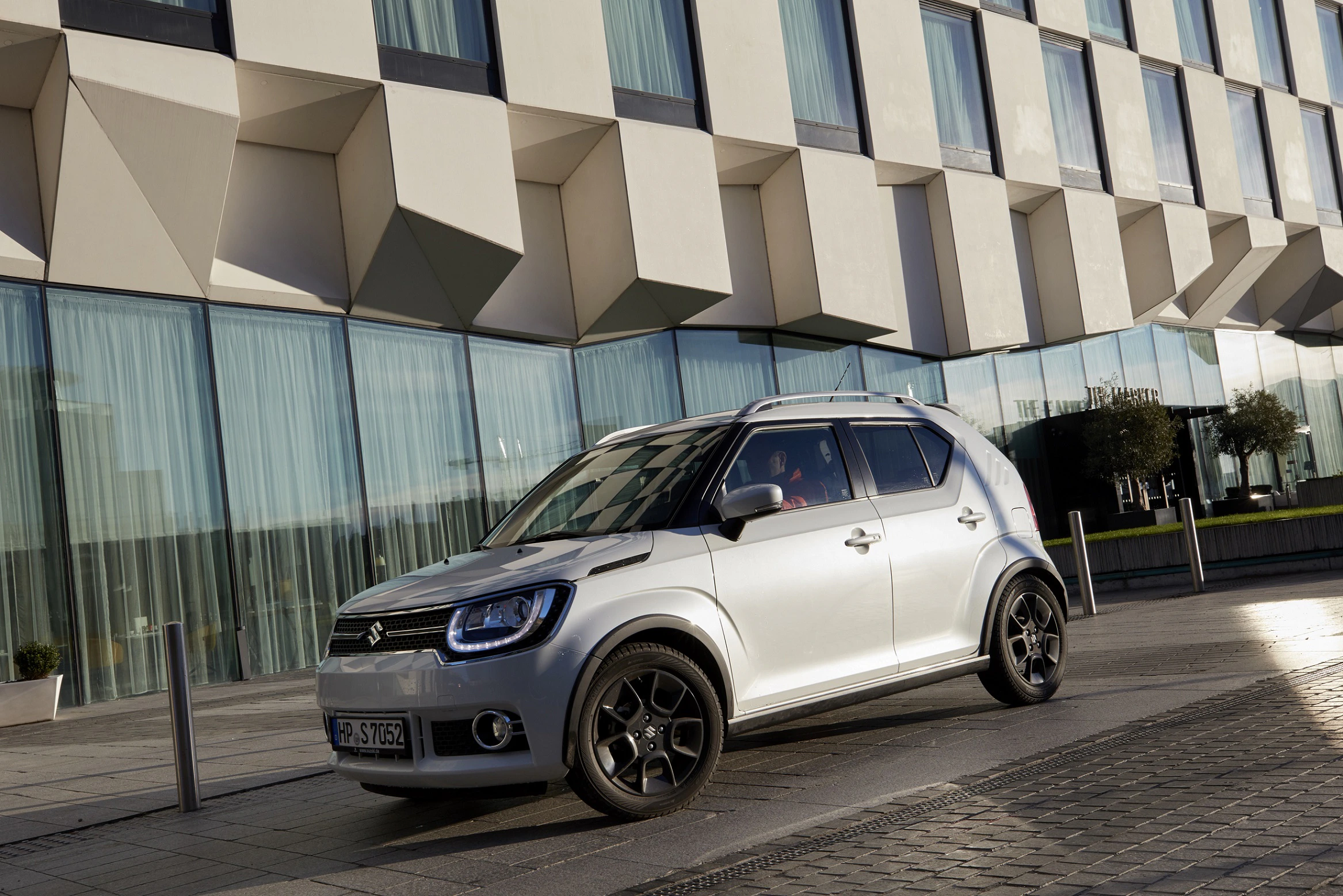 Kompakter silberner SUV parkt vor modernem Gebäude mit geometrischem Fassadendesign. Sonnenlicht erzeugt klare Schatten.