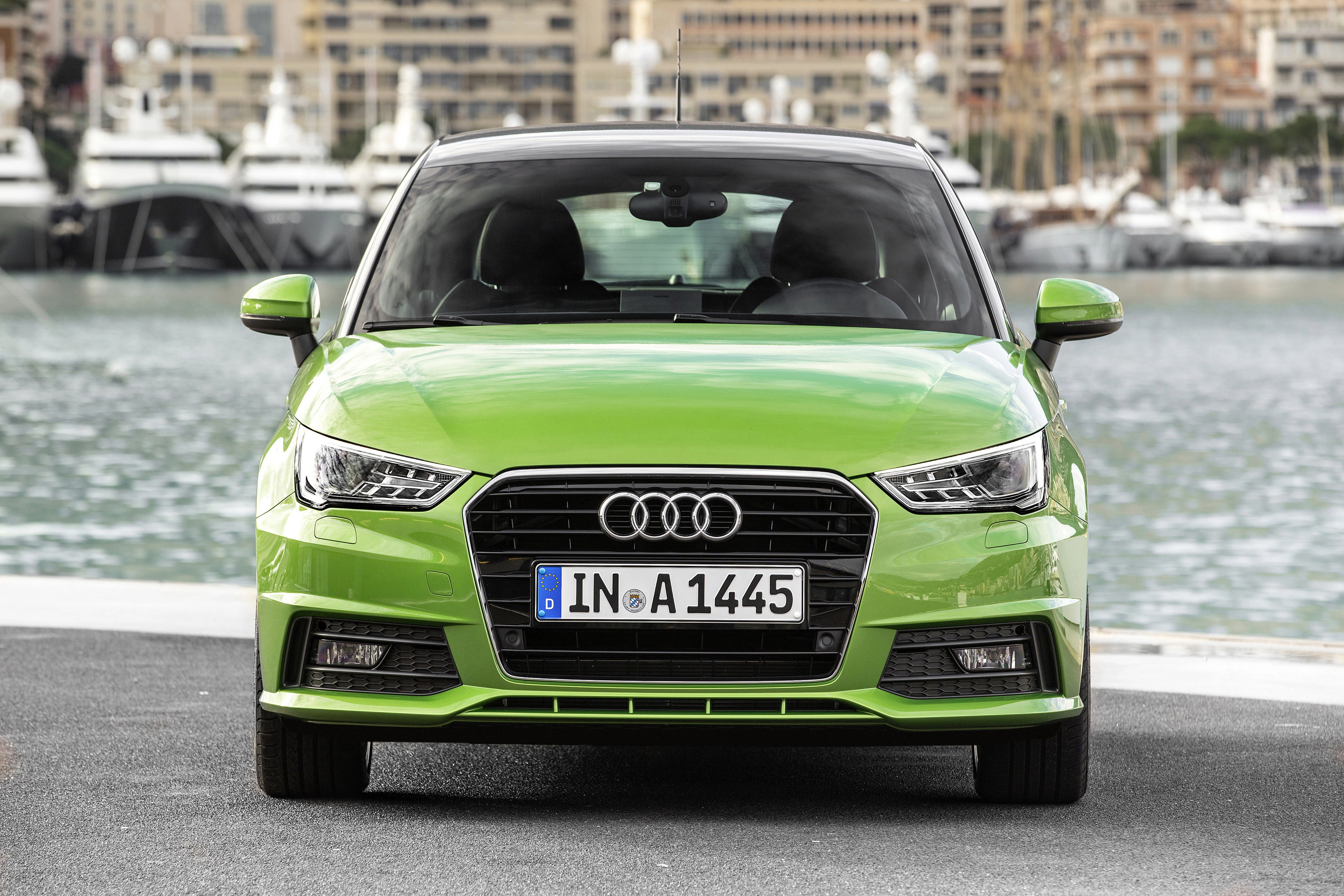 Grüner Audi A1 parkt vor Yachthafen, im Hintergrund sind Boote und Gebäude zu sehen. Vorderansicht des Autos mit deutschem Kennzeichen.