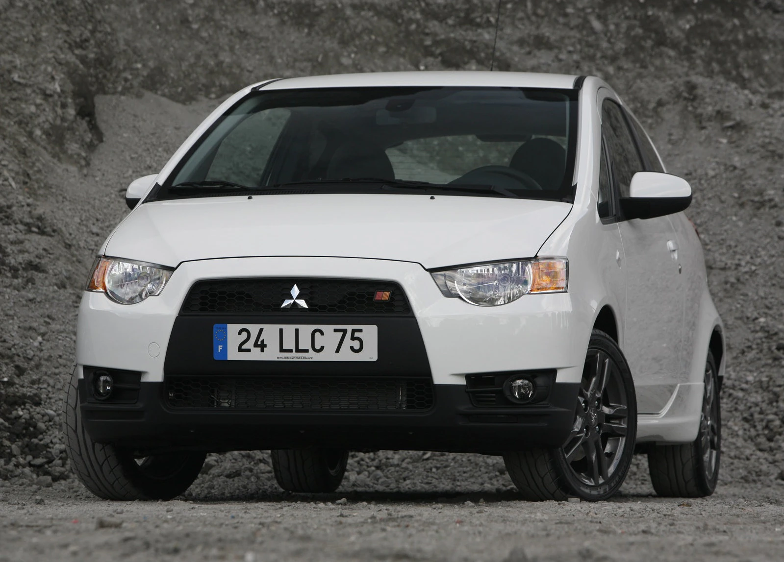 Weißer Mitsubishi Colt vor grauem Hintergrund, schwarze Felgen und sportliches Design. französisches Kennzeichen.