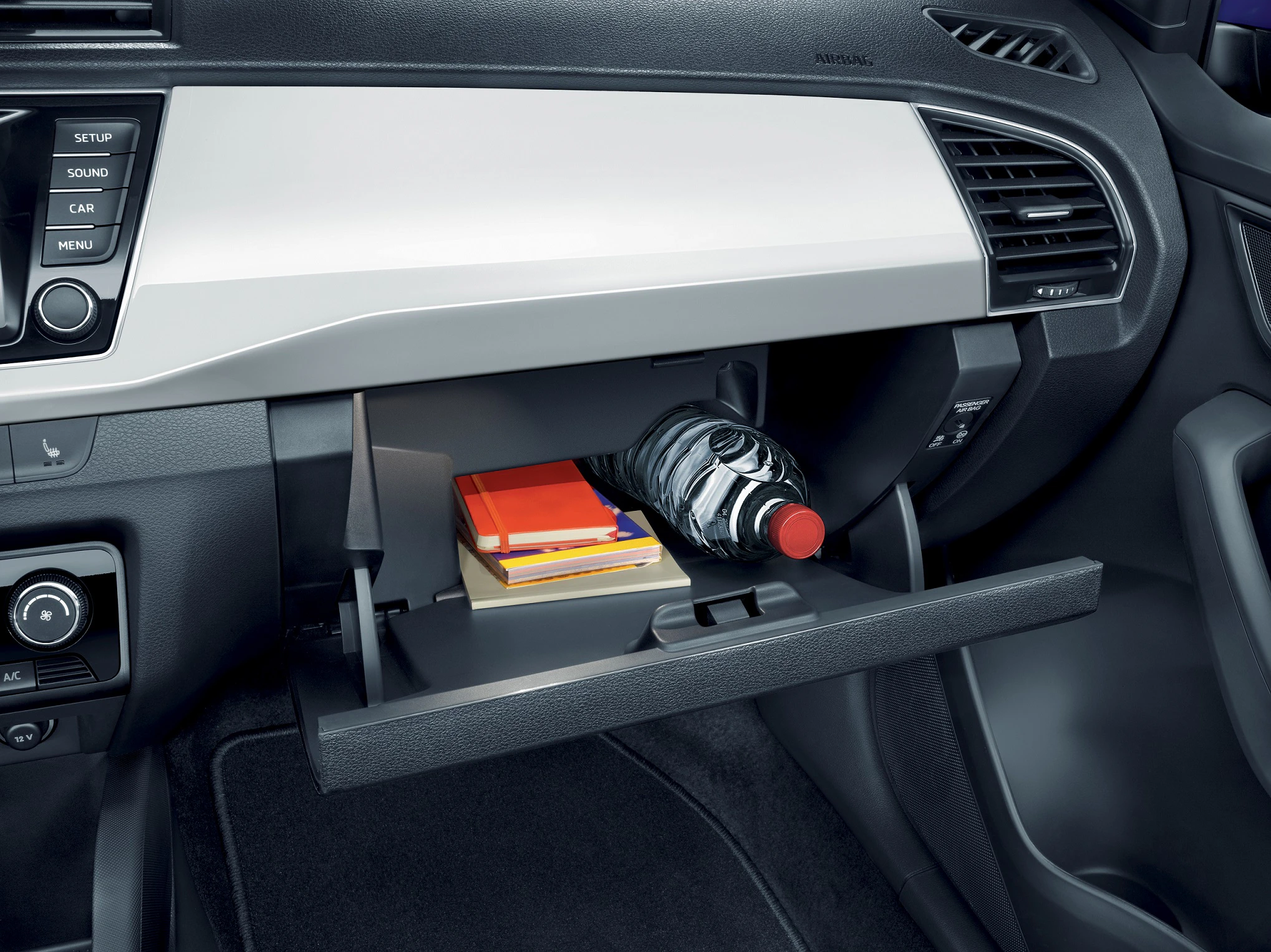 Ein offenes Handschuhfach eines Autos mit Büchern und einer Wasserflasche. Daneben Bedienelemente und Belüftungsöffnung.