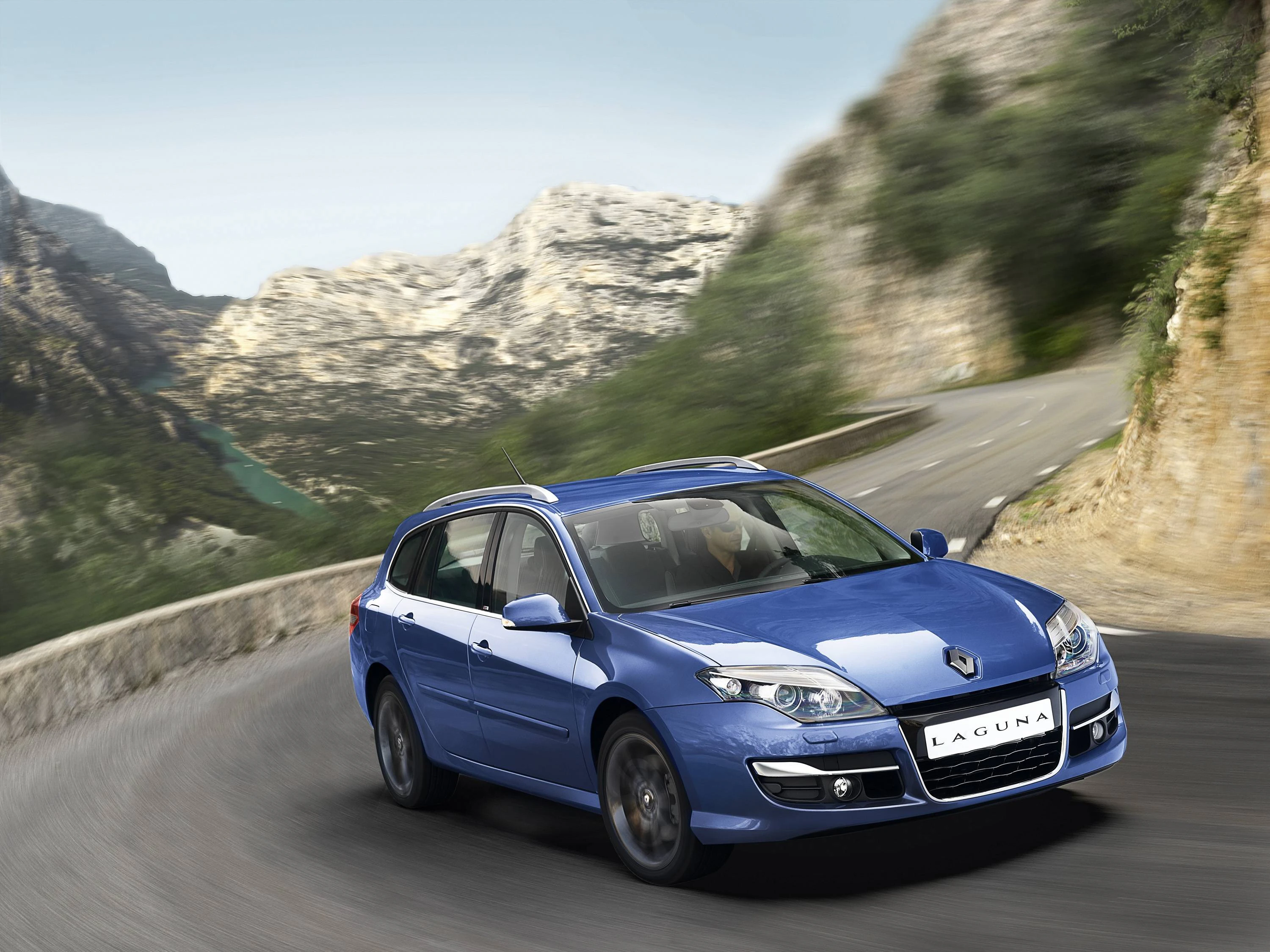 Blauer Renault Laguna fährt auf kurviger Bergstraße mit Felsen und bewaldeten Hängen im Hintergrund. Dynamische, malerische Szenerie.