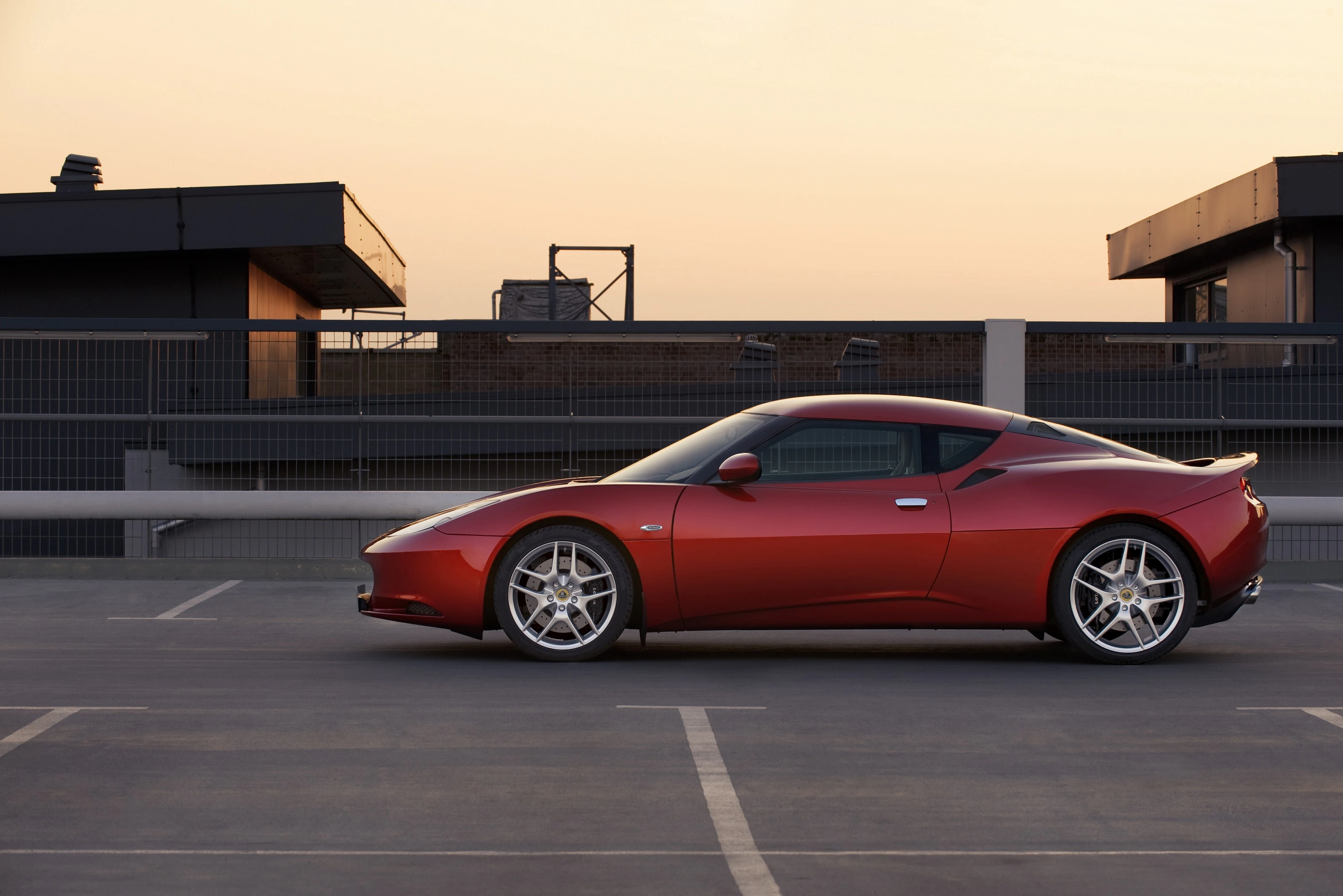Rotes Sportauto auf leerem Parkdeck bei Sonnenuntergang, Seitenansicht, mit Gebäuden im Hintergrund.