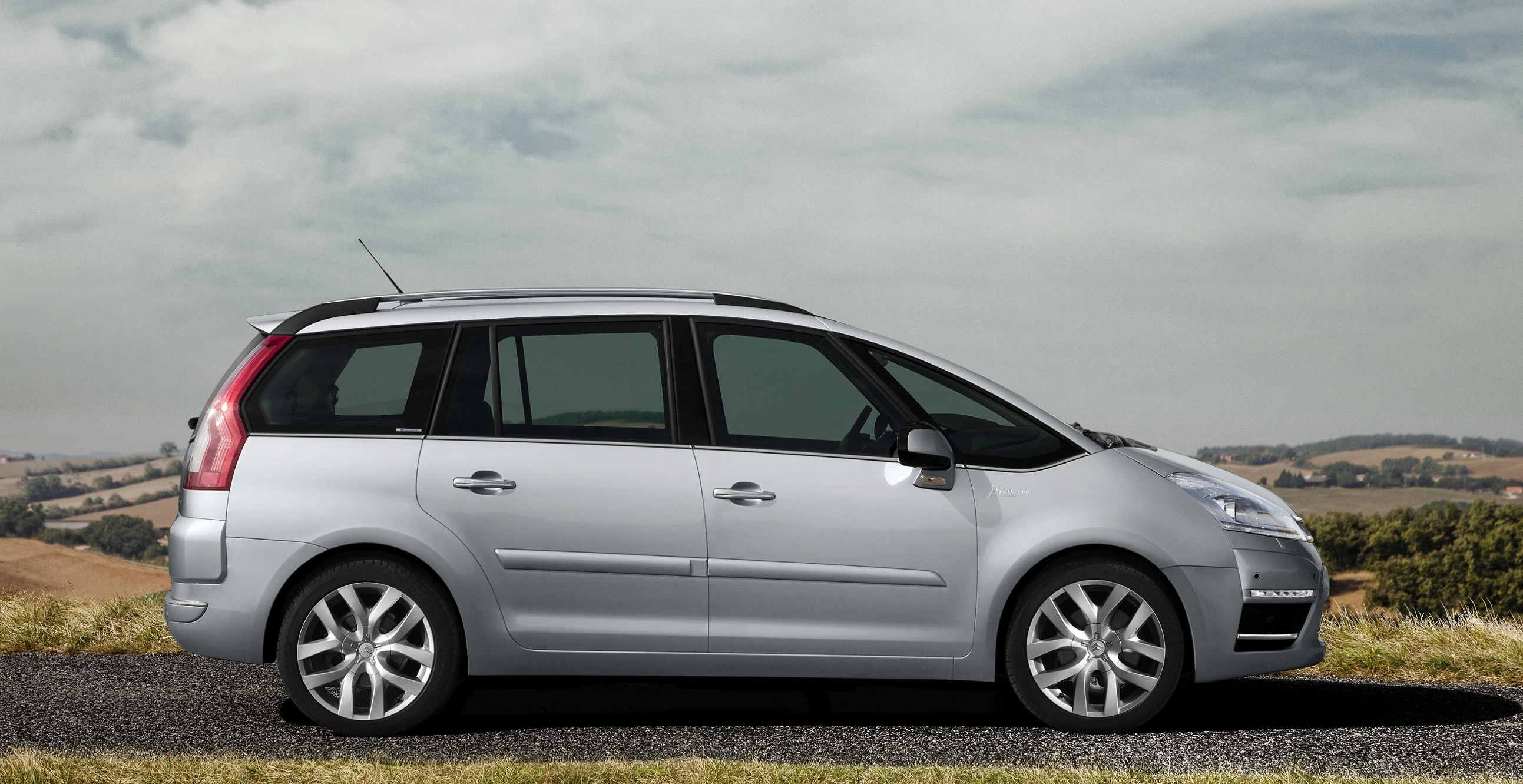 Silberner Minivan auf einem ländlichen Weg mit Wolkenhimmel im Hintergrund.
