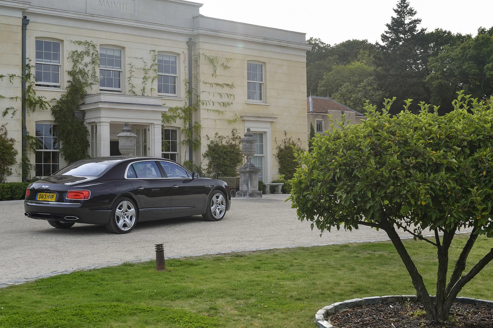 Schwarzer Luxuswagen parkt vor einem eleganten, mit Efeu bewachsenen Herrenhaus. Ein gepflegter Garten mit Bäumen umgibt das Gebäude.