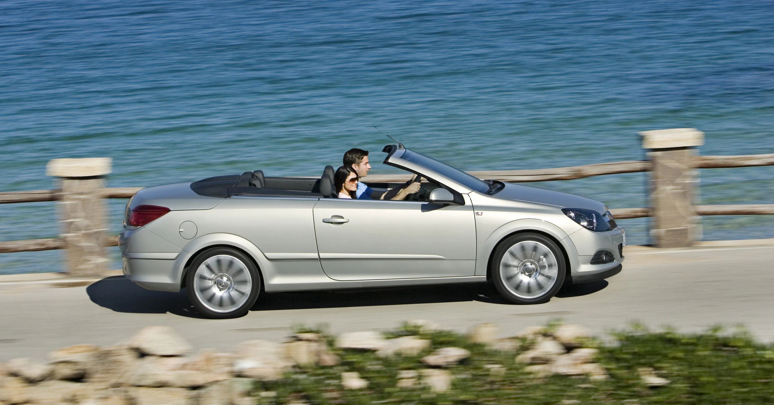 Silbernes Cabrio mit offenem Dach fährt auf einer Küstenstraße am Meer entlang. Die Sonne scheint und der Fahrer genießt die Aussicht.