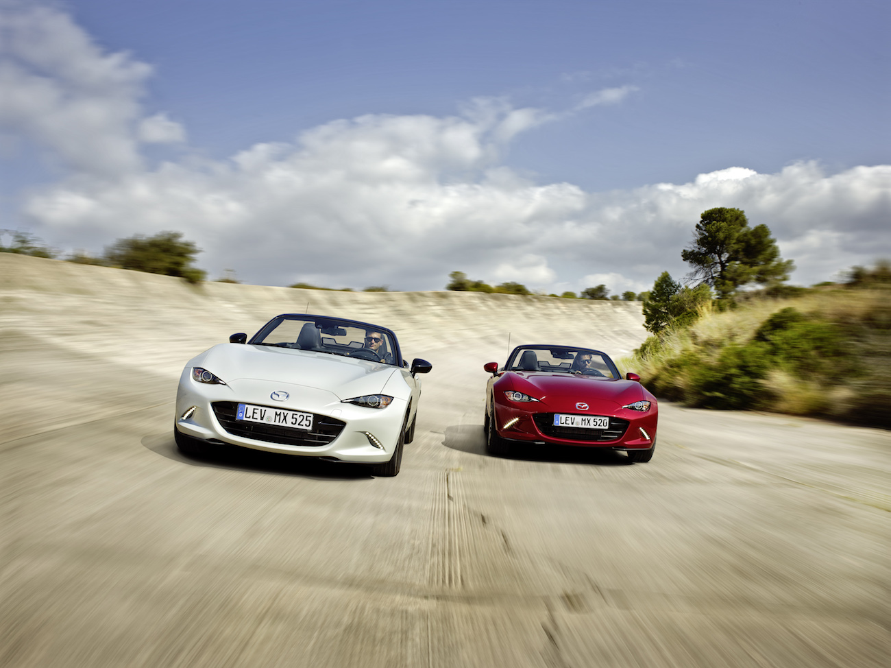 Zwei Mazda MX-5 Cabriolets in Weiß und Rot fahren nebeneinander auf einer Straße unter blauem Himmel mit Wolken.