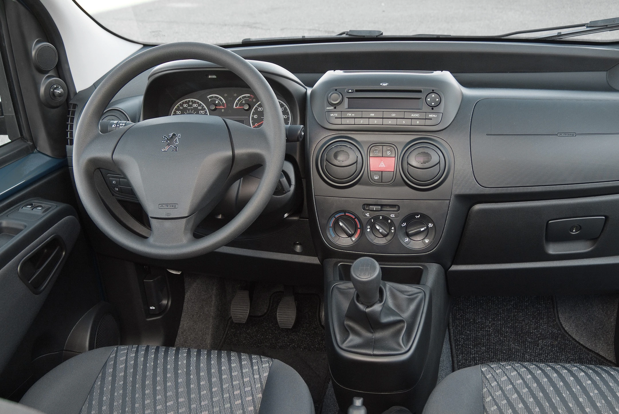 Innenraum eines Autos mit grauen Sitzen, Lenkrad, Schalthebel, Armaturenbrett und Bedienelementen; Fokus auf Fahrerkabine.