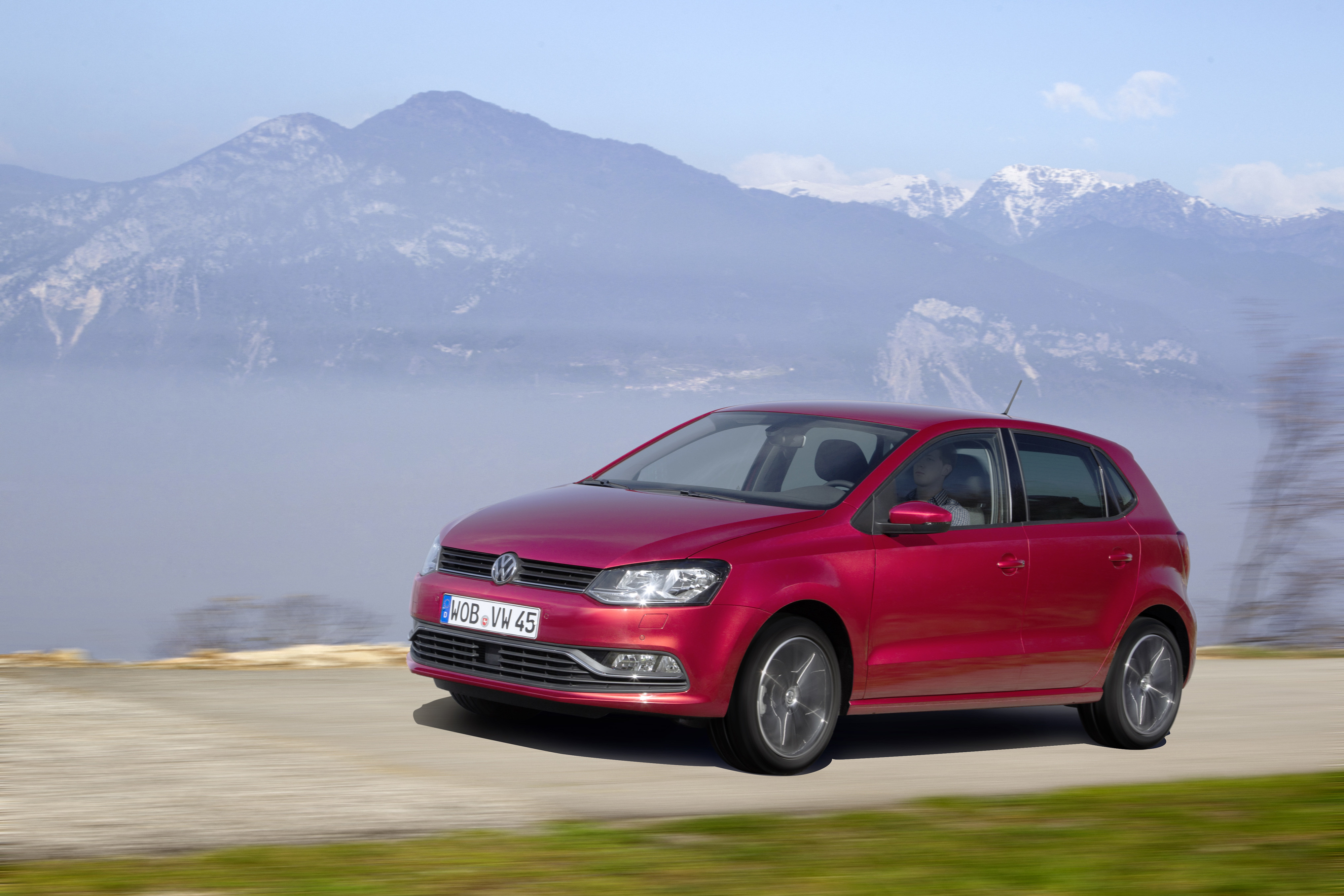 Roter VW Polo V mit Facelift-Front fährt vor Bergpanorama mit Schneegipfeln über eine Schotterstraße.